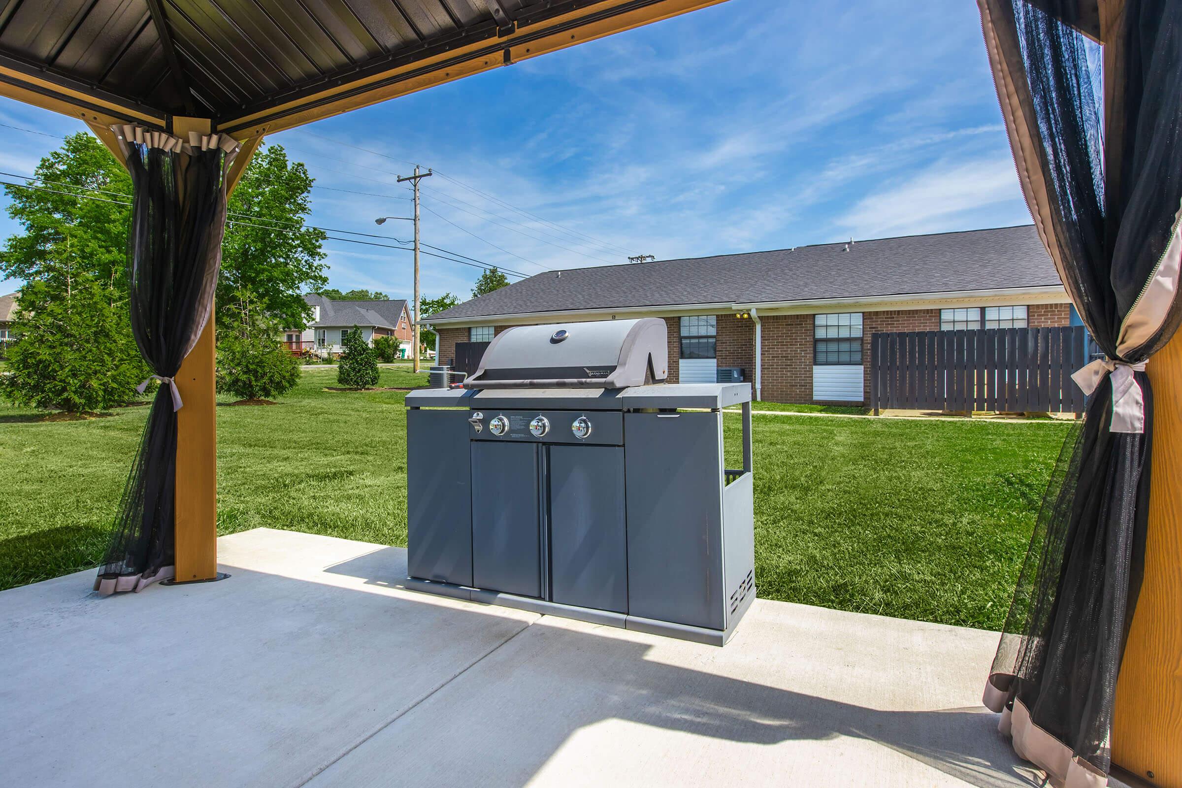 A covered outdoor patio featuring a modern grill, set against a backdrop of a clear blue sky and well-maintained green lawn. The scene includes a glimpse of neighboring homes in the distance.