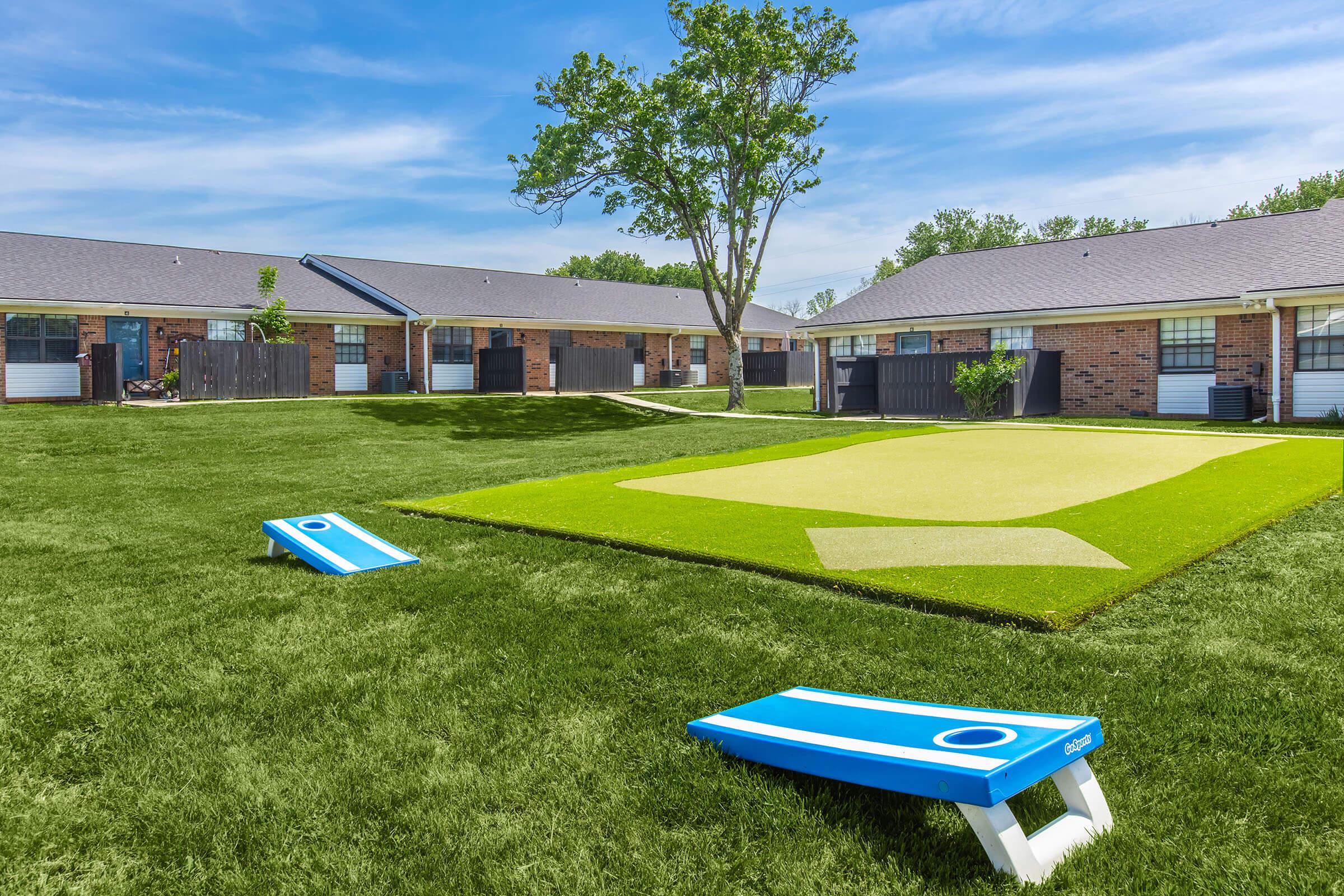 A sunny outdoor area featuring a well-maintained lawn with two cornhole boards on the left and a green turf surface resembling a putting green in the background. Surrounding the area are several single-story brick buildings, creating a relaxed atmosphere for recreational activities.