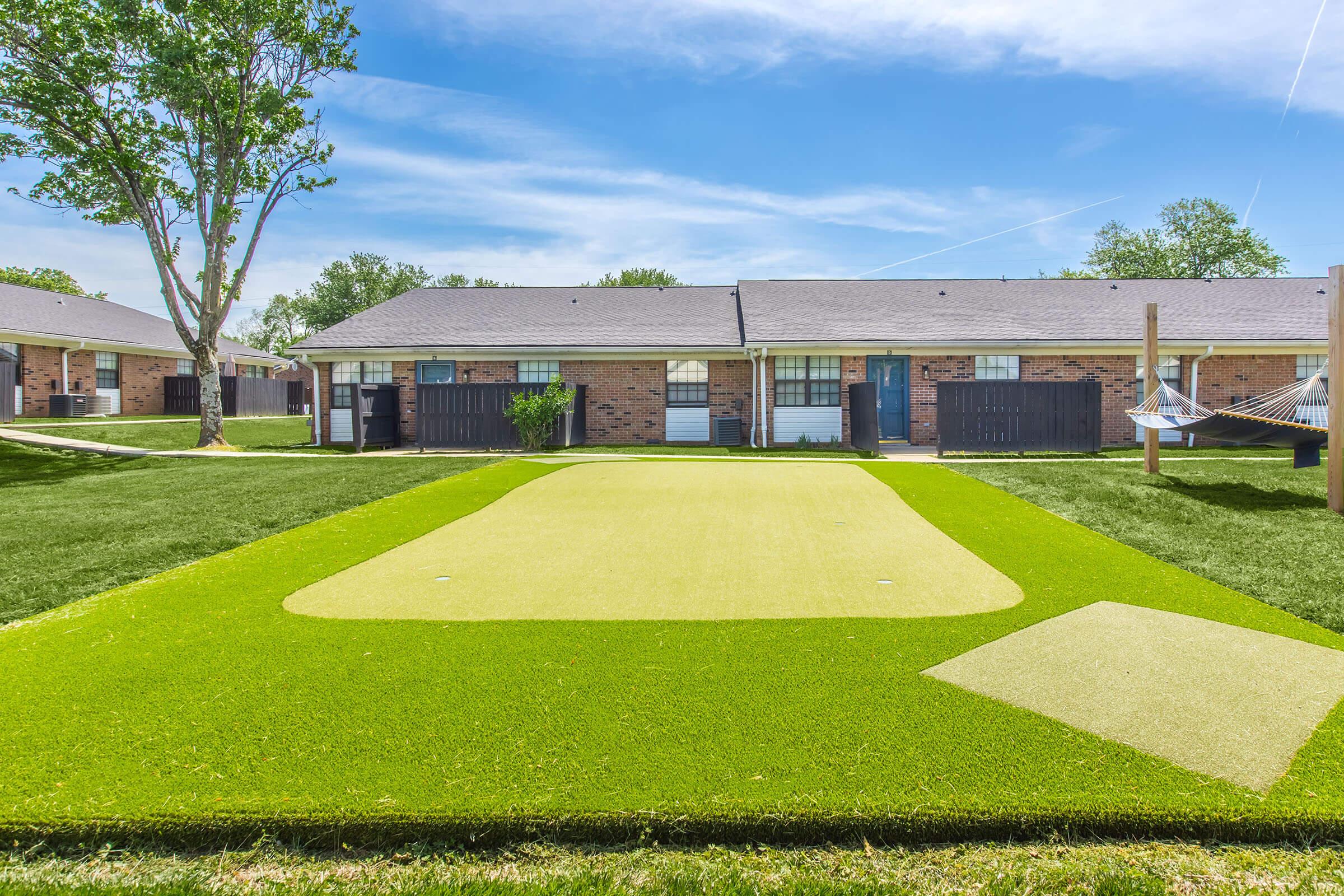 A grassy area featuring a putting green, with a small patch of turf on the side. In the background, there are brick buildings with windows and doors, surrounded by trees and a clear blue sky. A hammock is visible, adding to the relaxed outdoor atmosphere.