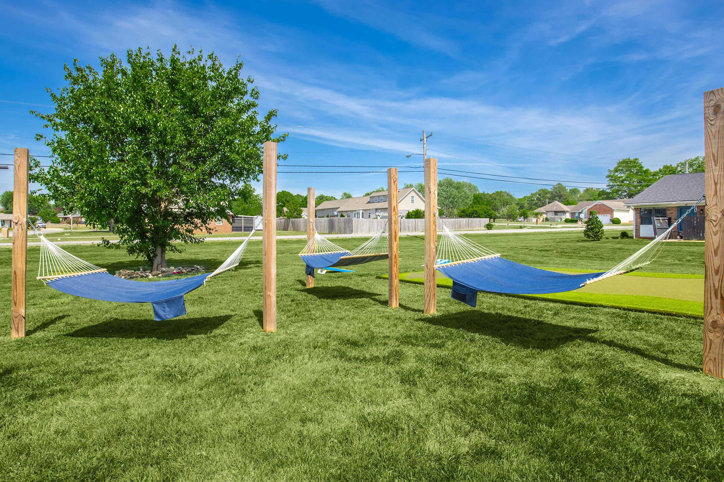 Three blue hammocks are strung between wooden posts in a grassy park. A vibrant green tree stands nearby, and houses are visible in the background under a clear blue sky. The scene conveys a feeling of relaxation and tranquility.