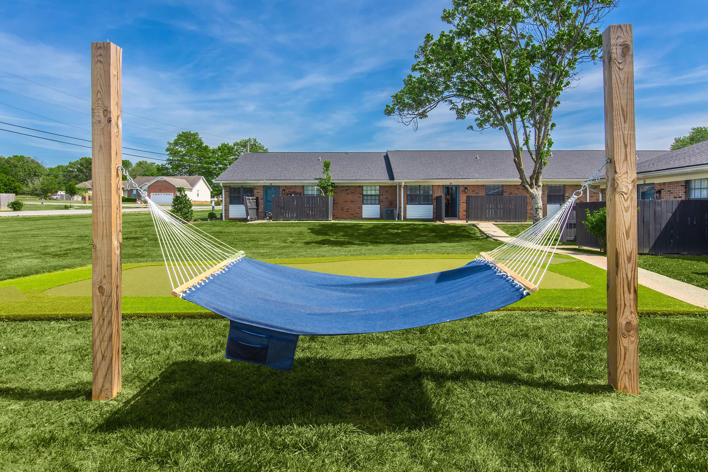 A blue hammock is suspended between two wooden posts, set in a green grassy area. In the background, there are several single-story buildings and trees under a clear blue sky. The scene is peaceful and inviting, ideal for relaxation.