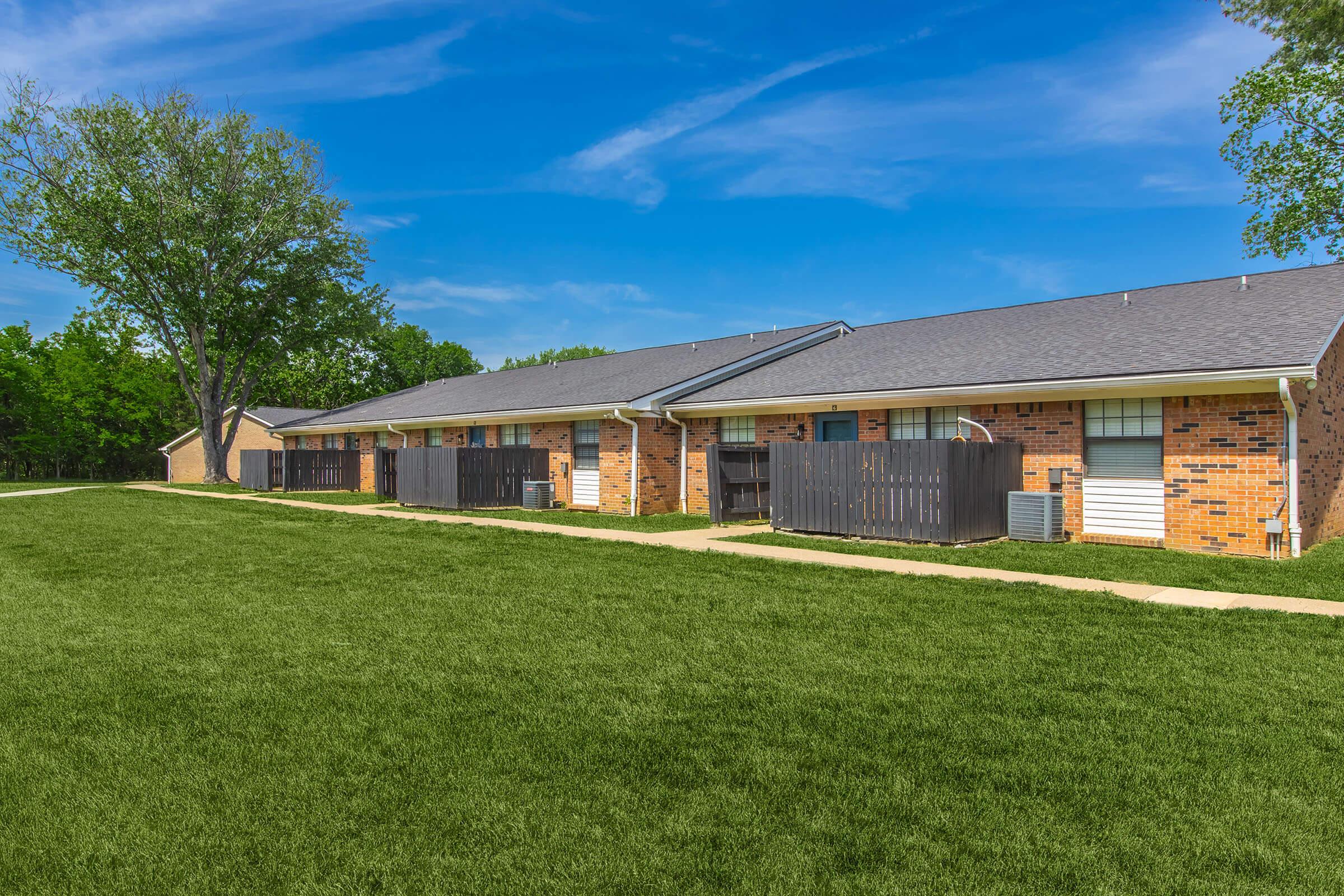 A row of brick apartment buildings with dark roofs, set against a bright blue sky. Each unit has a small fenced patio and lawn in front. Green grass covers the ground, and trees are seen in the background, giving a pleasant, residential atmosphere.