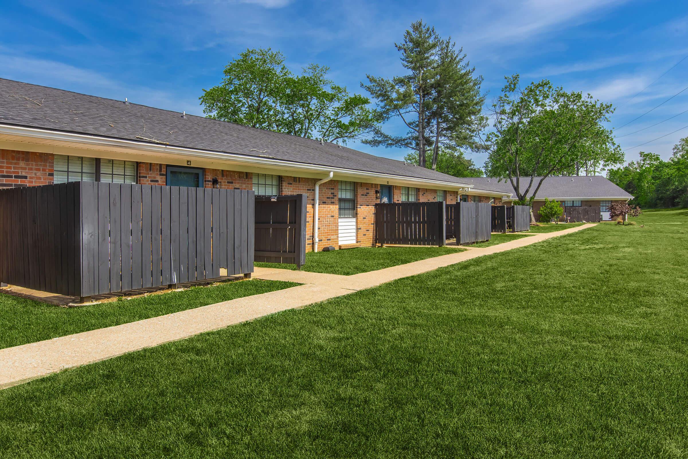 A row of residential buildings with brick exteriors and dark wooden fences, surrounded by well-maintained green grass. The pathway leads through the property, lined with trees and a clear blue sky in the background.