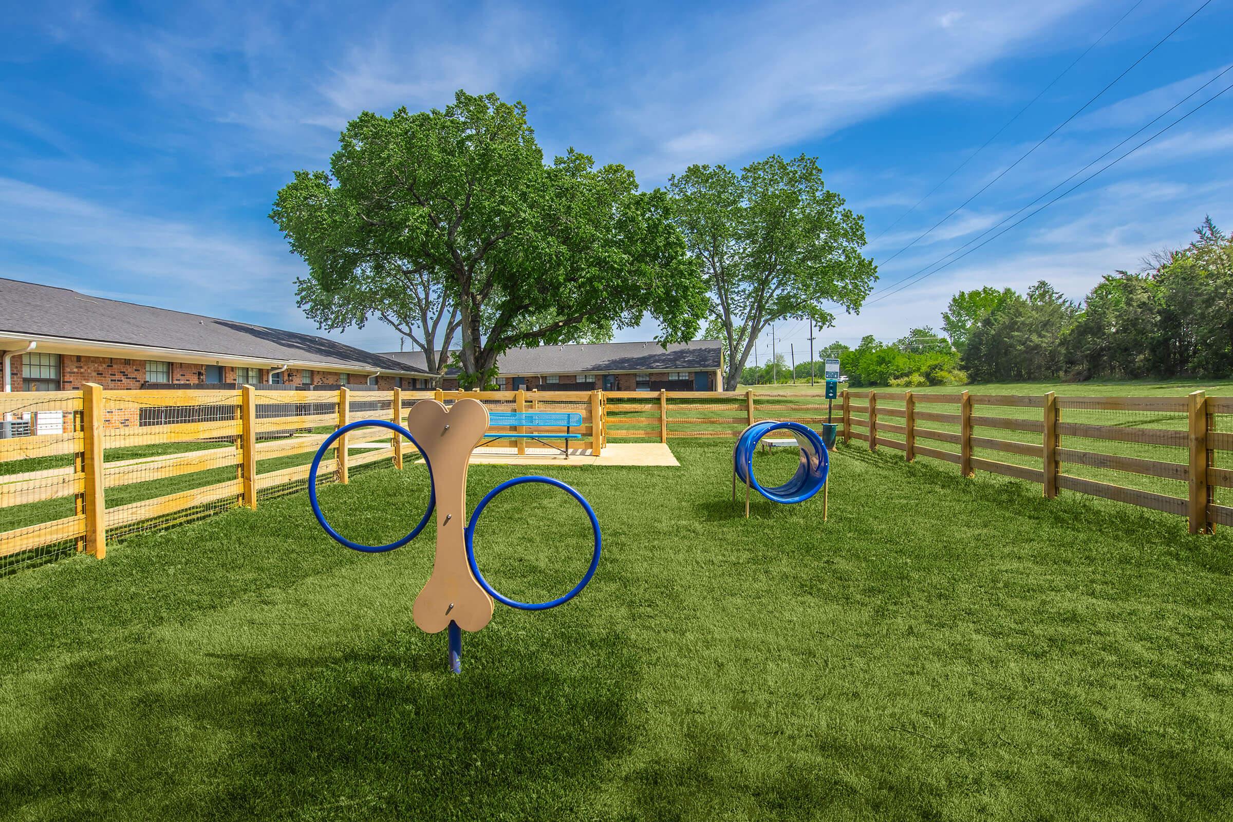 A well-maintained outdoor dog park featuring green grass, wooden fencing, and various agility equipment, including a large bone-shaped structure and hoops. Trees provide shade, and there is a clear blue sky overhead, creating a welcoming and vibrant environment for dogs to play.