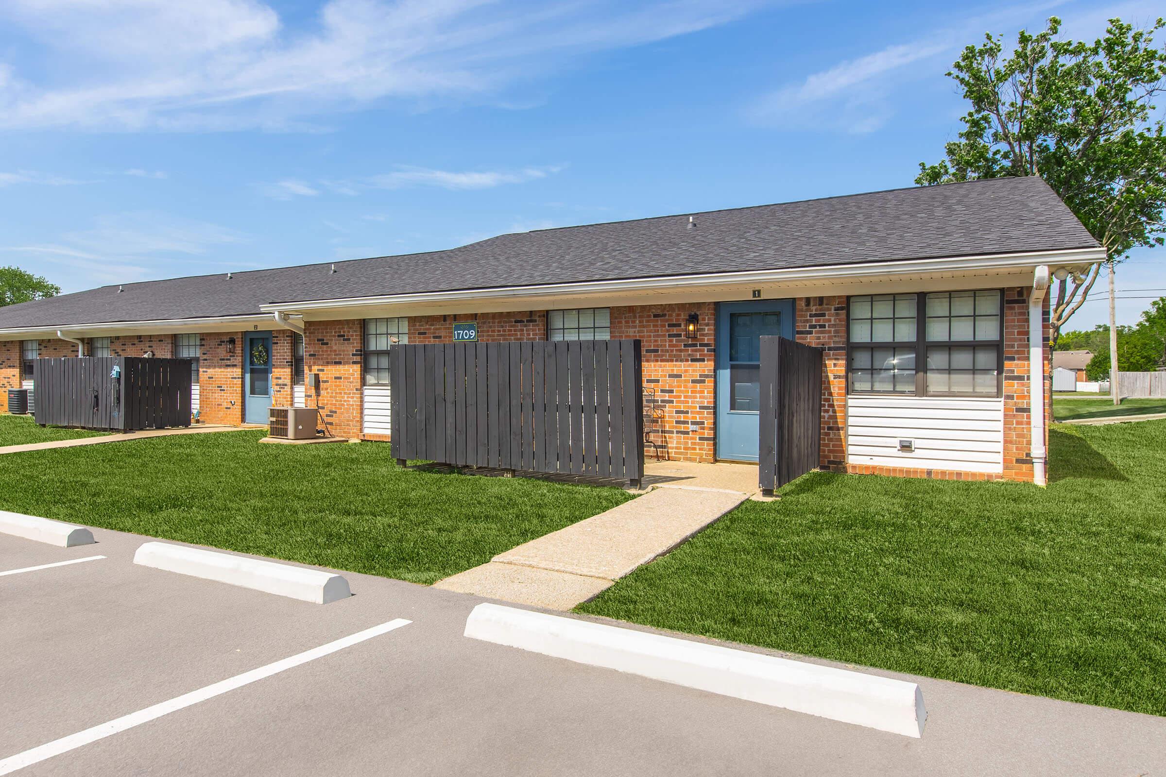 Single-story apartment building with brick facade, featuring two units separated by a wooden fence. Each unit has a front door with a porch area and grassy landscaping in front. The parking lot is visible with marked spaces, and there are trees and a blue sky in the background.