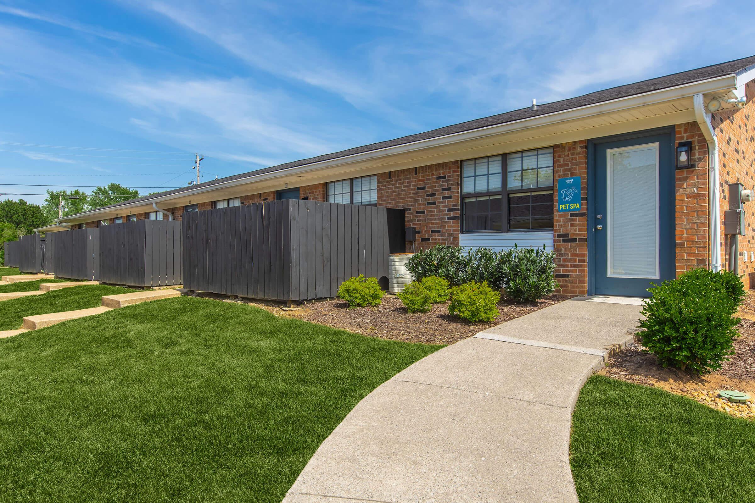 A well-maintained brick building with a green lawn and neatly trimmed bushes. A paved walkway leads to the entrance, which has a dark door and a property sign. Wooden fences are visible, providing privacy to the outdoor area. The sky is clear with a few clouds, indicating a sunny day.