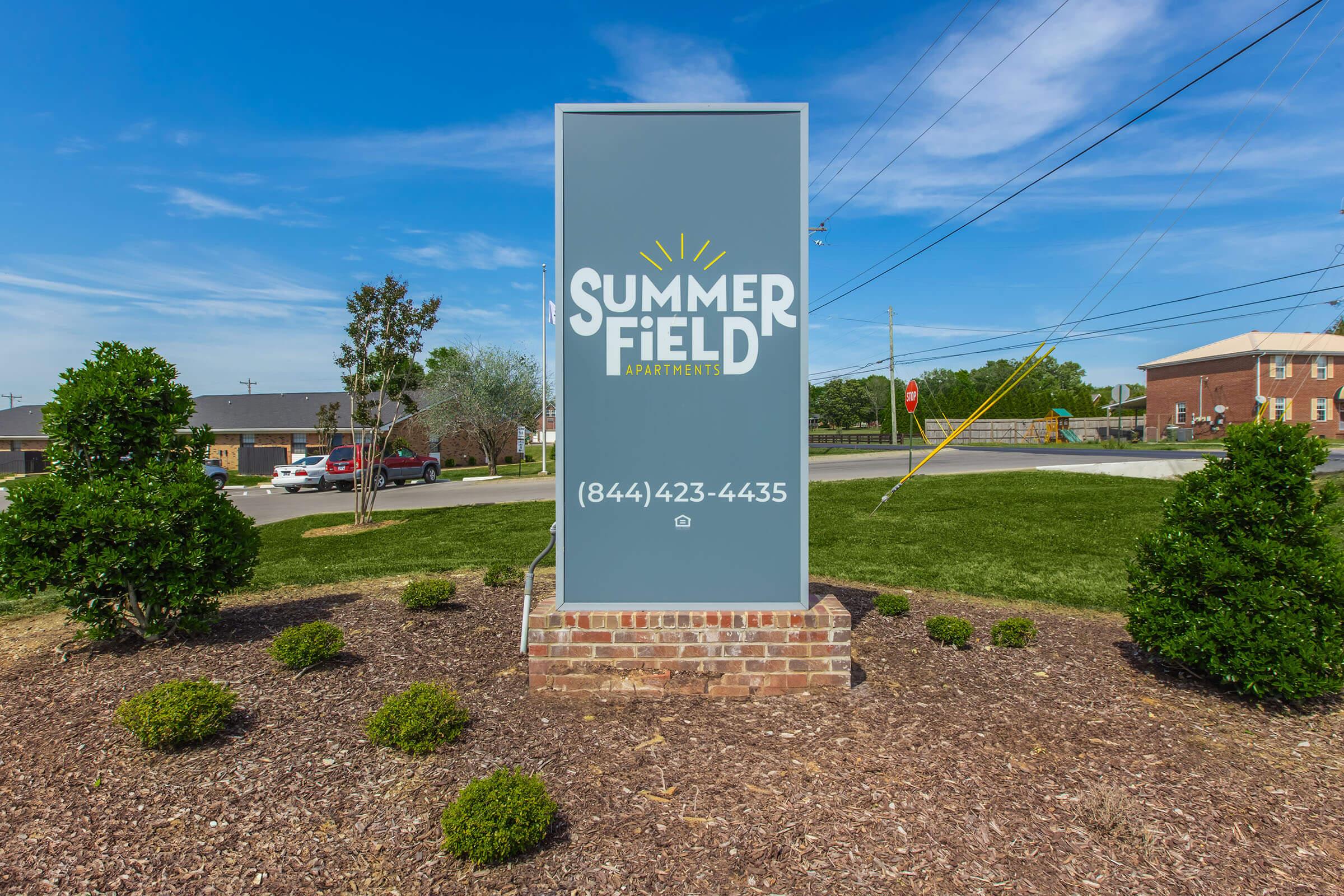 Sign for Summer Field Apartments displaying the name "SUMMER FIELD" in large letters with a sun graphic above. The contact number is (844) 423-4435. The sign is placed on a landscaped area with small bushes and grass, set against a clear blue sky.