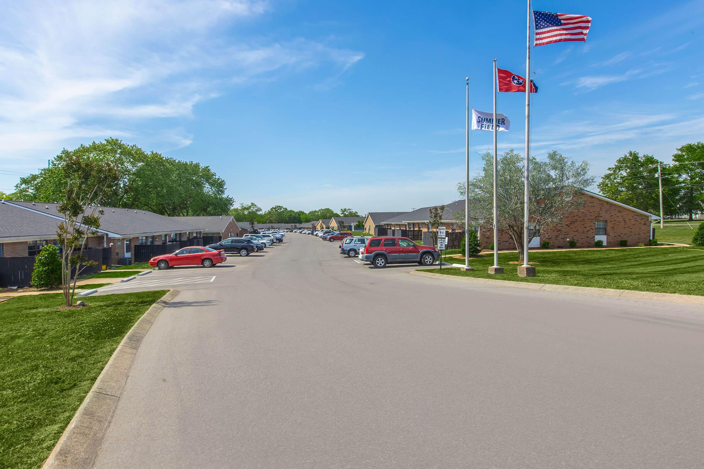 A wide view of a residential street lined with parked cars on both sides. In the foreground, there are two flagpoles displaying the American flag and a state flag. On either side, brick buildings with lawns are visible, indicating a well-maintained neighborhood under a clear blue sky.