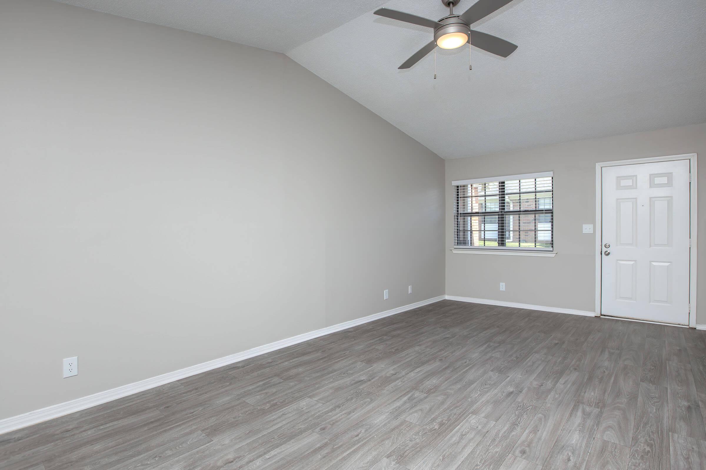 Empty room with light gray walls and a vaulted ceiling. The flooring is a wood-like laminate. A ceiling fan is mounted in the center, and there is a window with blinds on one wall, as well as a white door leading outside. Natural light brightens the space.