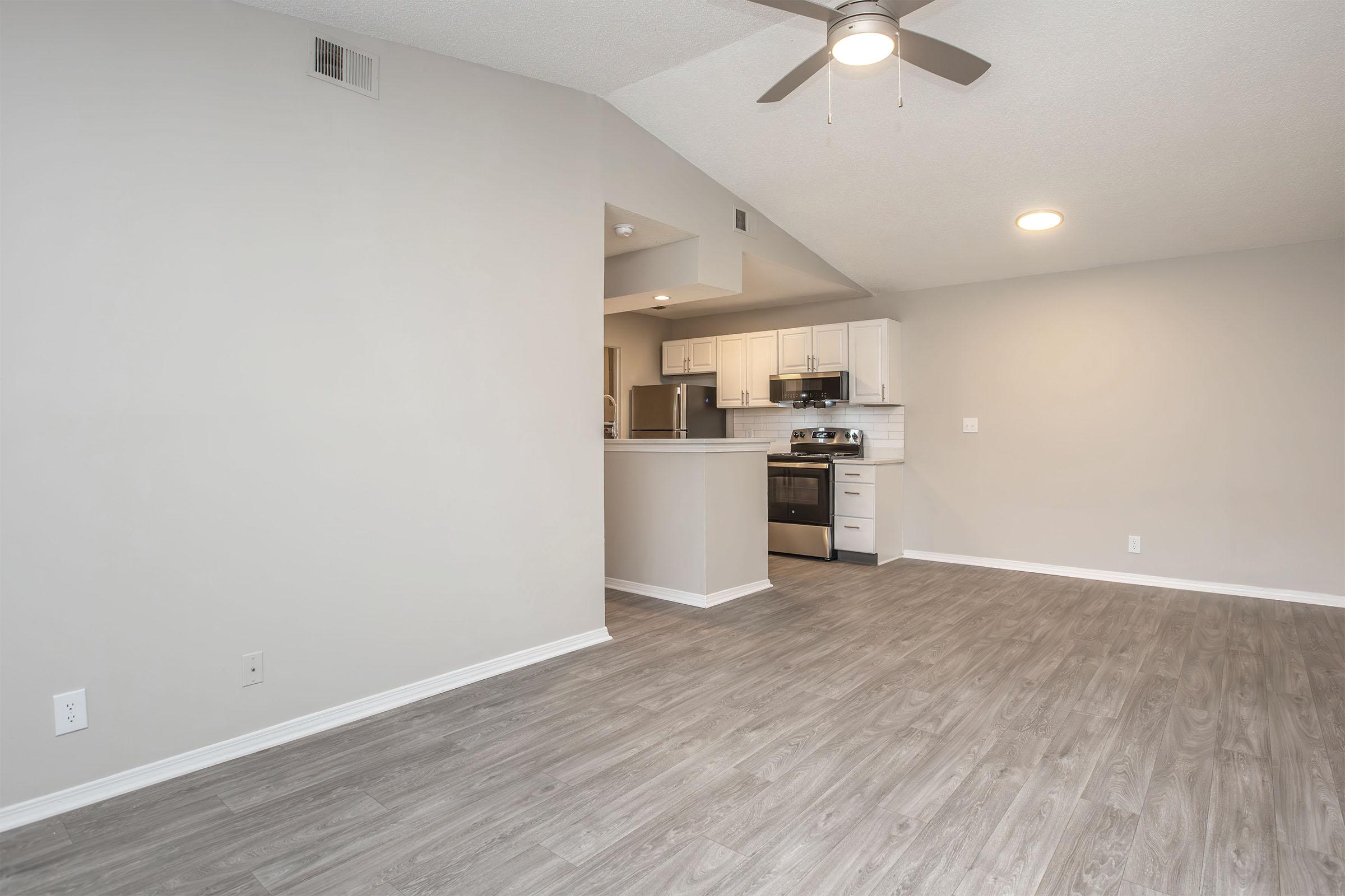 Interior of a modern apartment living space with gray walls and laminate flooring. The open-concept area features a ceiling fan and leads to a kitchen with white cabinets and stainless steel appliances. Natural light comes in from a window, enhancing the airy feel of the room.