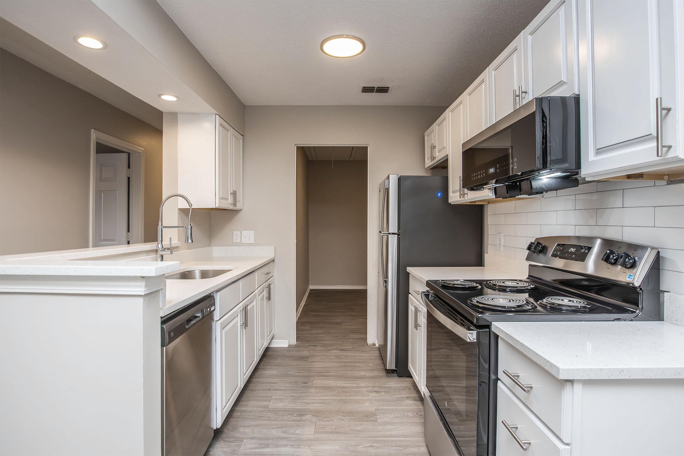 A modern kitchen featuring white cabinets, stainless steel appliances, and a spacious layout. The kitchen includes a double sink, dishwasher, and an electric stove, complemented by light-colored countertops and a sleek backsplash. The floor is finished with a wood-like laminate, contributing to a clean and contemporary look.
