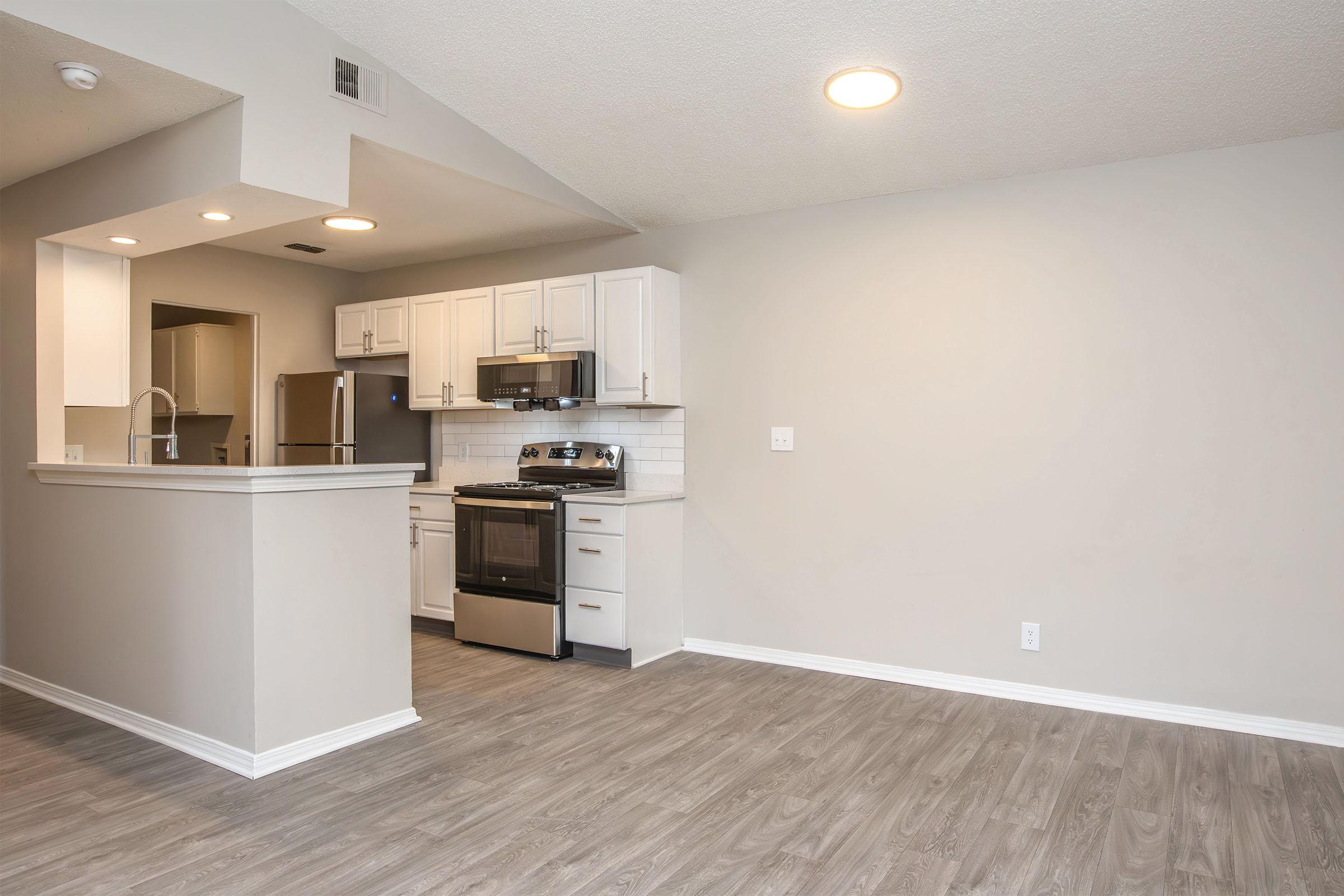 A modern kitchen with white cabinetry and stainless steel appliances, including an oven and refrigerator. The kitchen features a gray backsplash and an open layout, leading to a spacious living area with light wood-like flooring and neutral-colored walls. Two ceiling lights illuminate the space.