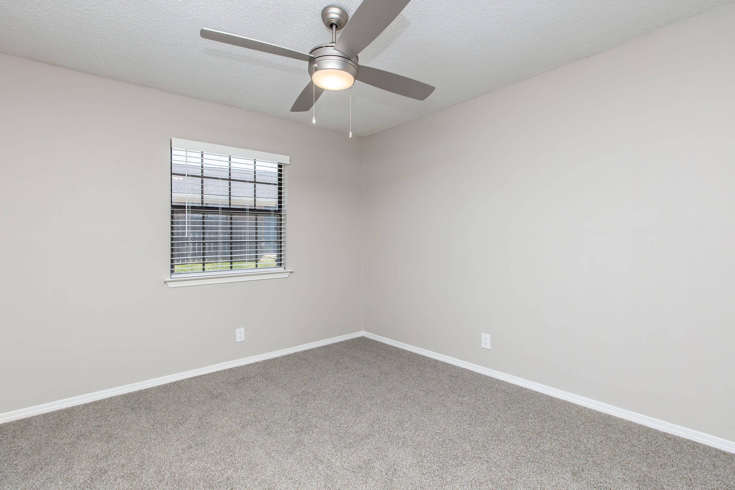 Empty room with light gray walls and beige carpet. A ceiling fan with light is centered on the ceiling. A window with blinds allows natural light, revealing a view of the outdoors. The room is unfurnished, creating a spacious and inviting atmosphere.