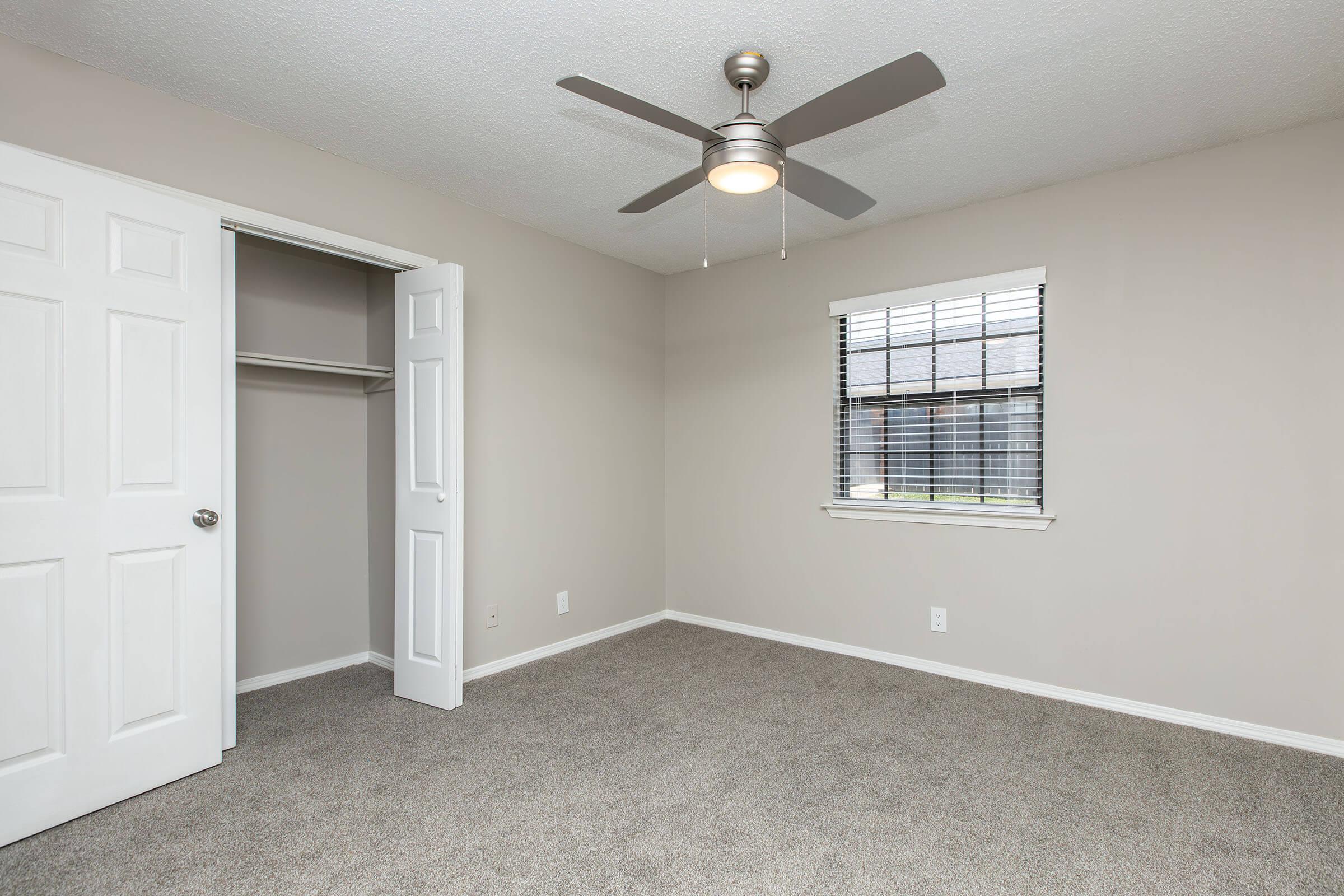 A well-lit, empty room with light beige walls and carpeted flooring. It features a ceiling fan, a window with blinds, and a closet with white double doors. The room has a clean and minimalist appearance, making it suitable for various uses.