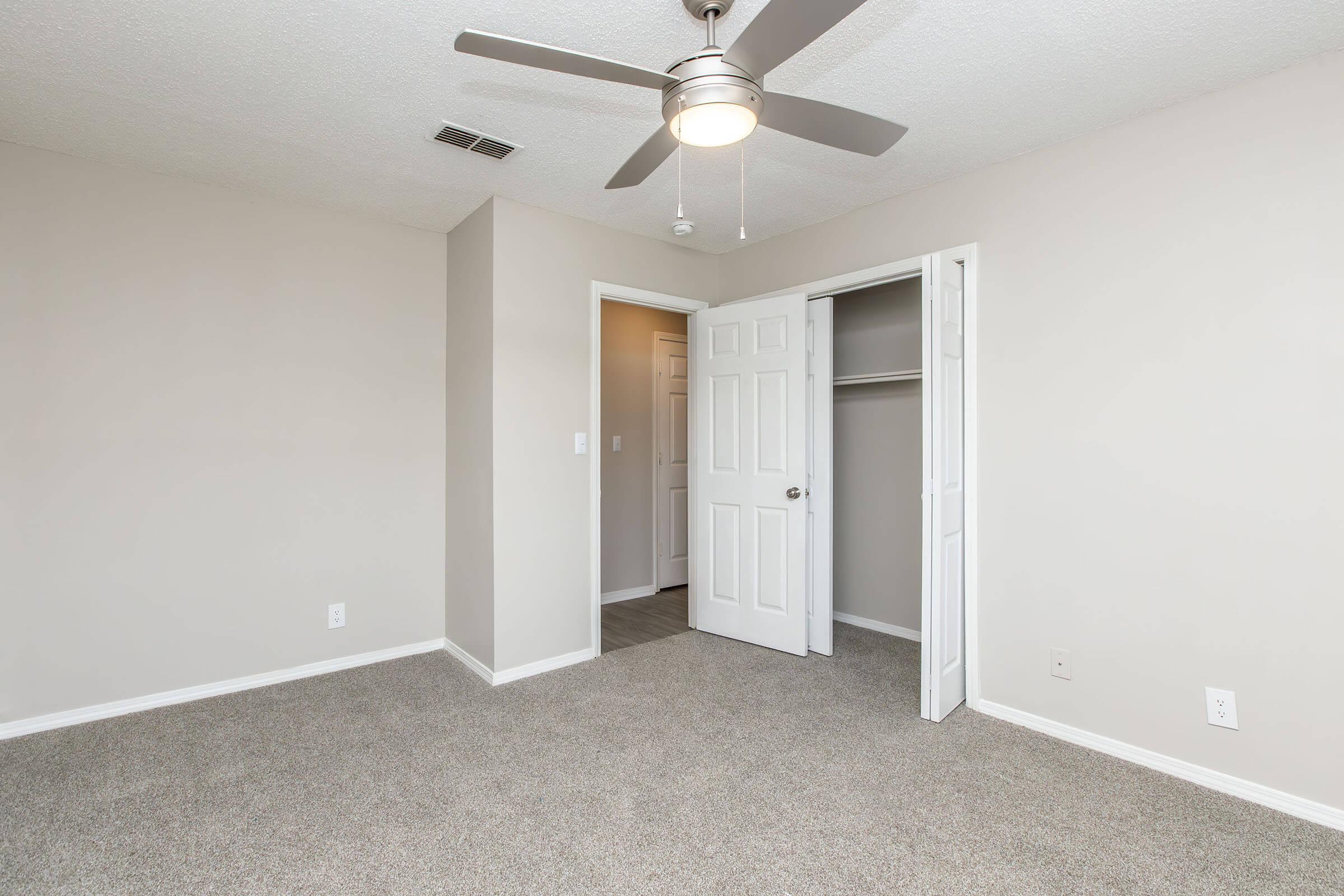 A neutral-toned bedroom featuring a ceiling fan, light gray carpet, and a white closet with double doors. The walls are painted light beige, and there is an open doorway leading to another room. The space is empty, creating a bright and airy feel.