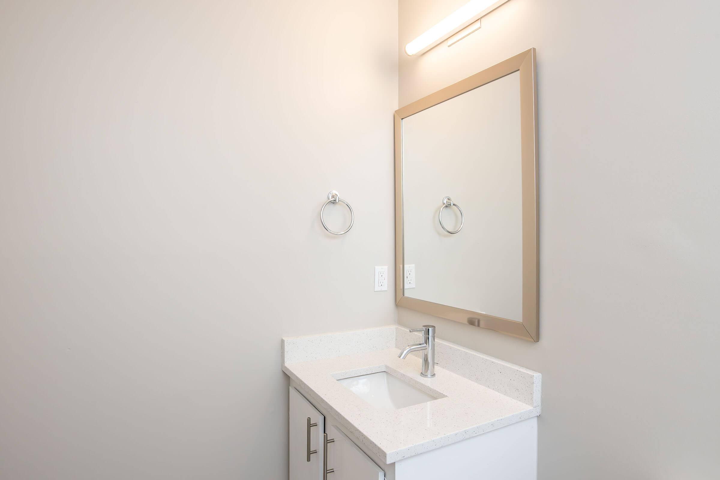 A modern bathroom with a minimalist design featuring a white countertop and a built-in sink. There is a large rectangular mirror with a simple frame mounted above the sink. A towel ring is mounted on the wall beside the mirror, and a light fixture is installed above the mirror, providing illumination. The wall is a neutral color.