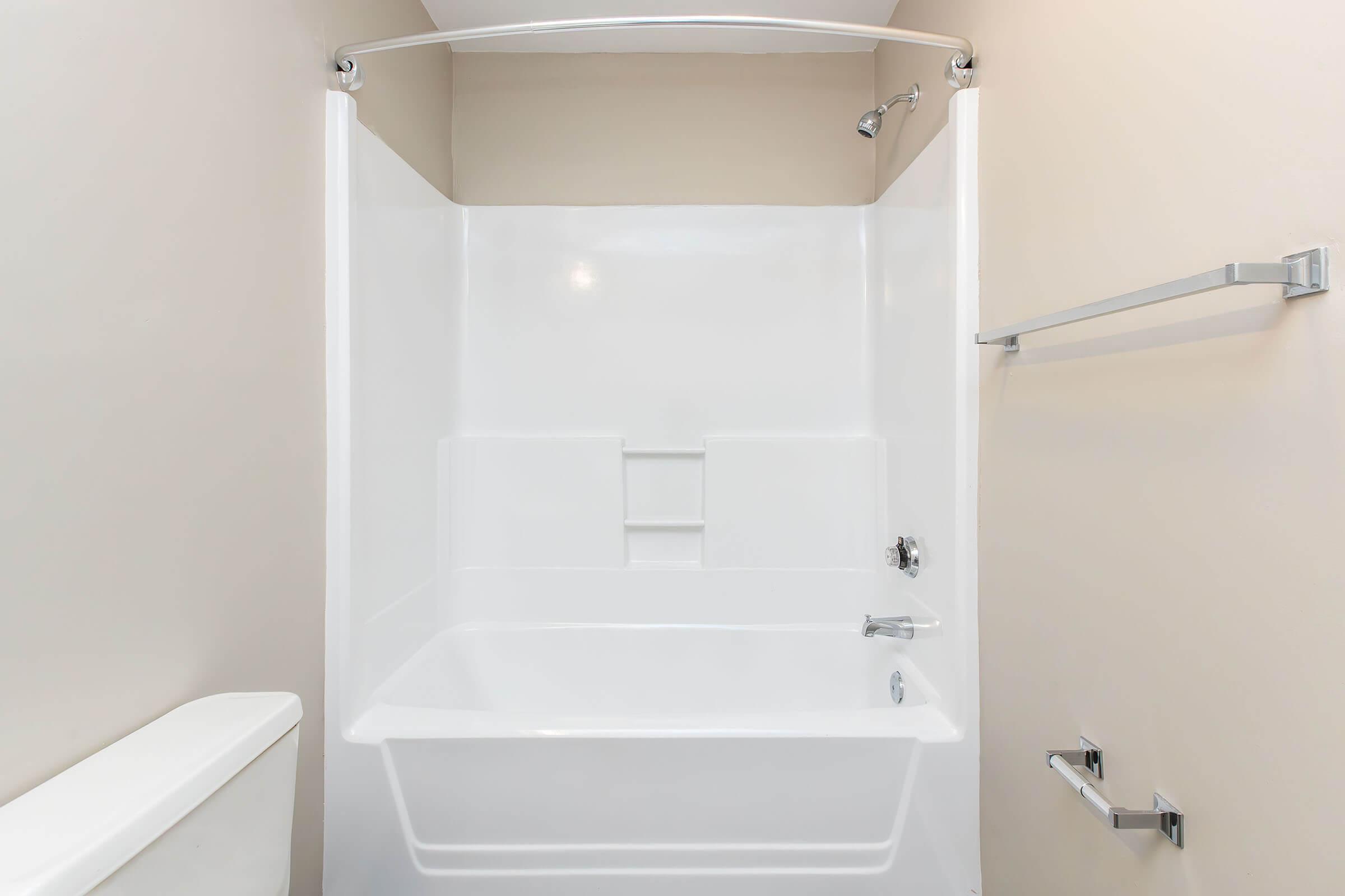 A clean, modern bathroom with a white bathtub and shower combination. The shower area features a curved shower rod and a single showerhead. The walls are a neutral beige color, and the bathroom includes a toilet and towel rail, creating a simple and functional space.