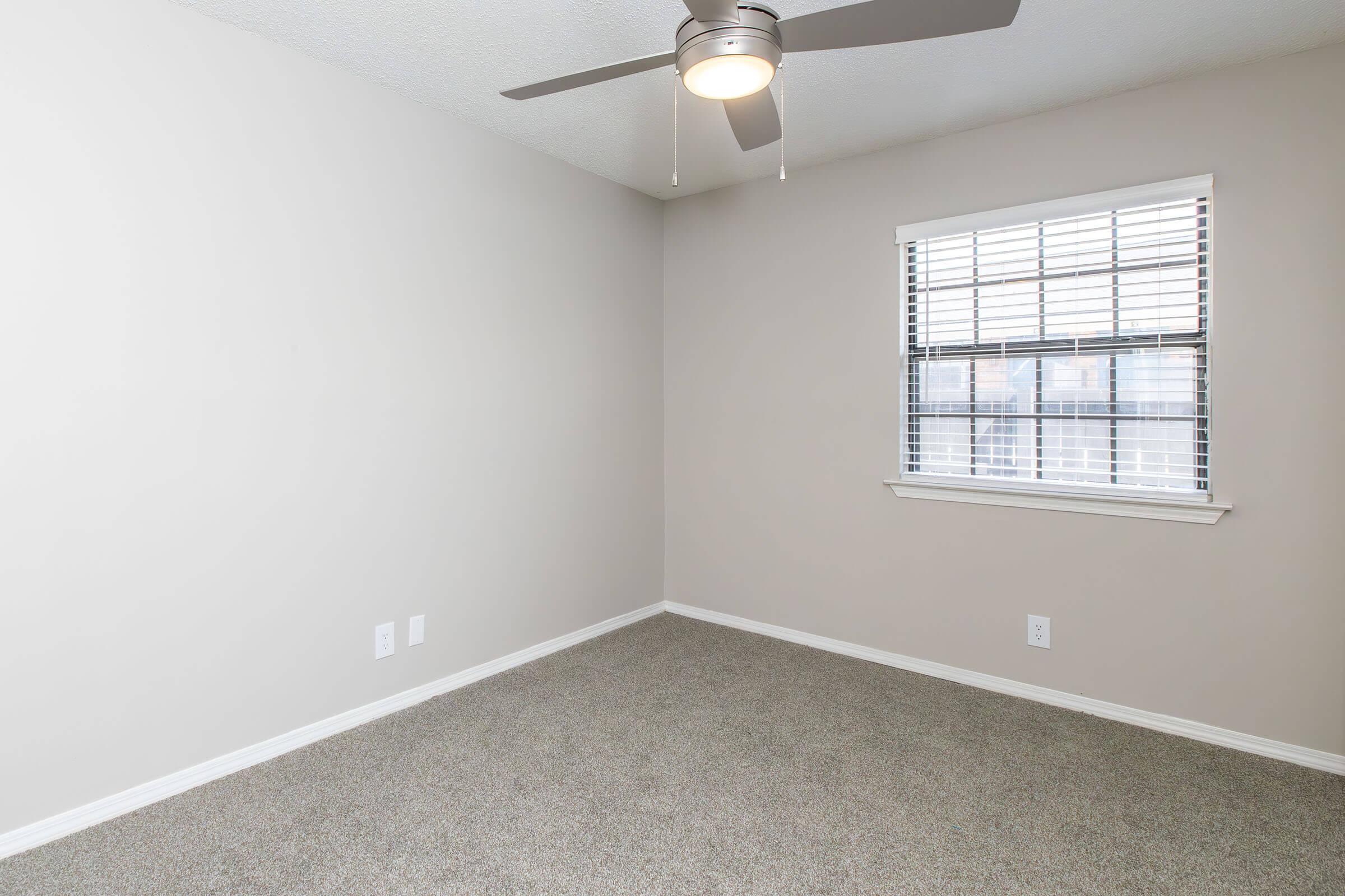 Empty room with light gray walls and carpeted flooring. A ceiling fan is mounted in the center, and a window with white blinds allows natural light to enter. The space is uncluttered, with two electrical outlets visible on one wall.
