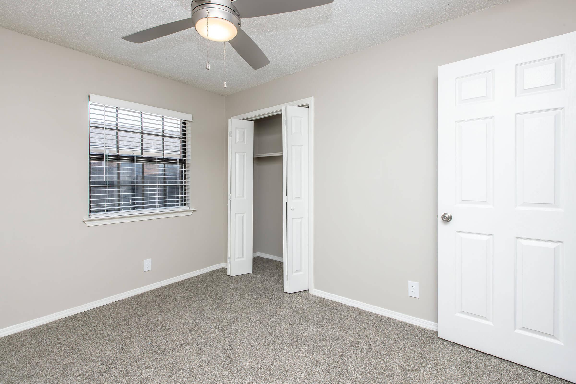 Empty bedroom featuring light gray walls, a ceiling fan, and a window with blinds. The carpet is also light gray. A closet with double doors is visible on the right, and a white, six-panel door leads out of the room. The overall atmosphere is bright and clean.