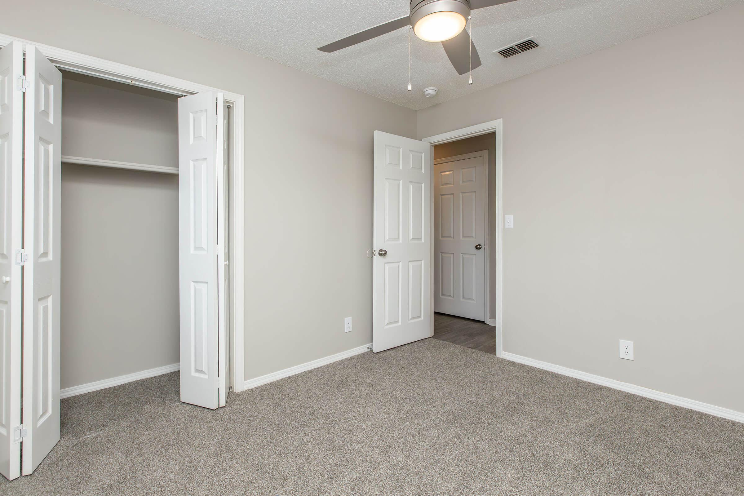 Empty room with beige walls and carpeted floor. Two open white doors lead to an adjoining room and a closet. A ceiling fan is visible above, and the room appears well-lit and spacious.