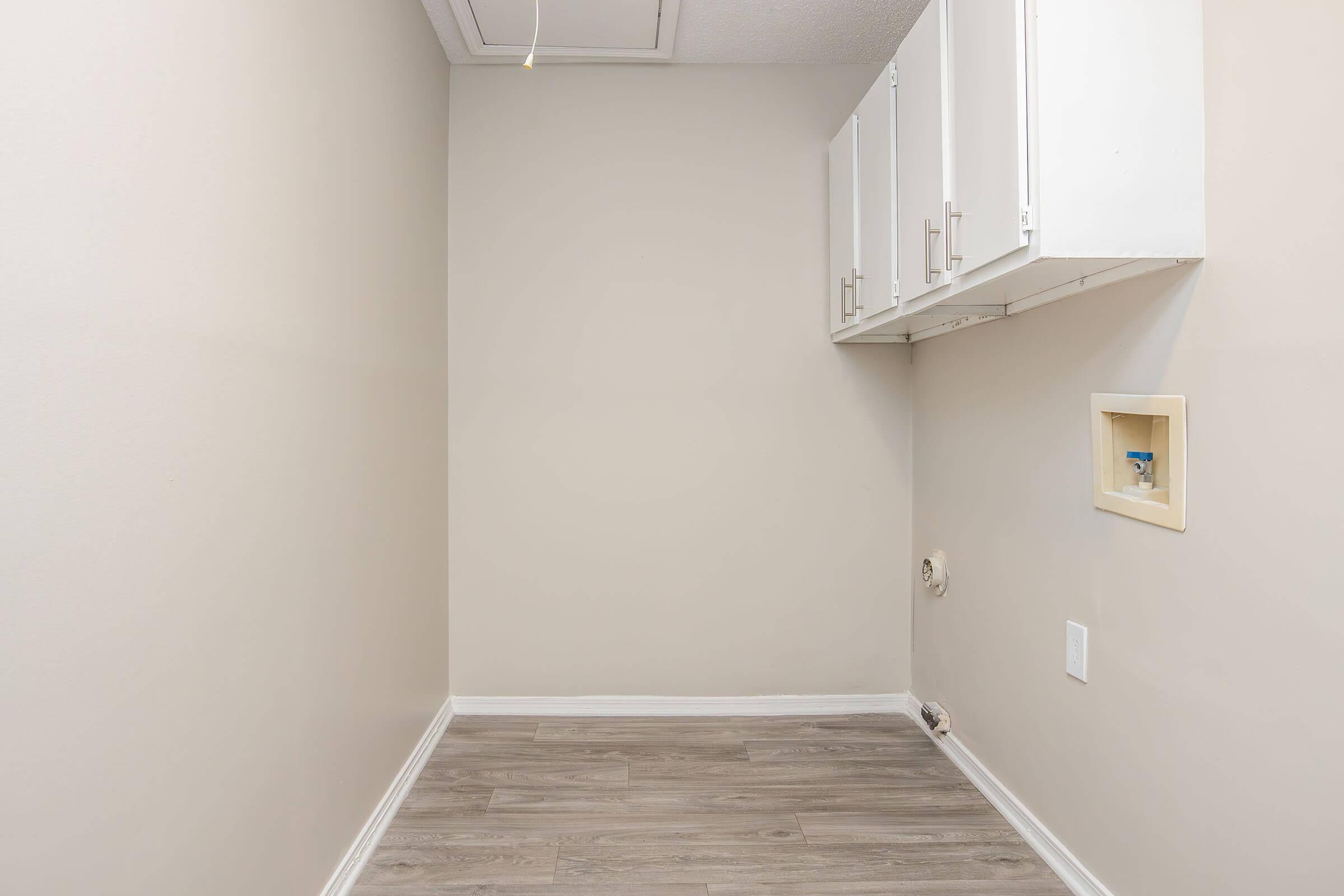 A spacious, empty laundry room featuring light beige walls, light wood flooring, and white cabinetry above. There is a small utility cutout in the wall and no appliances or furnishings present, creating a clean and minimalistic appearance.