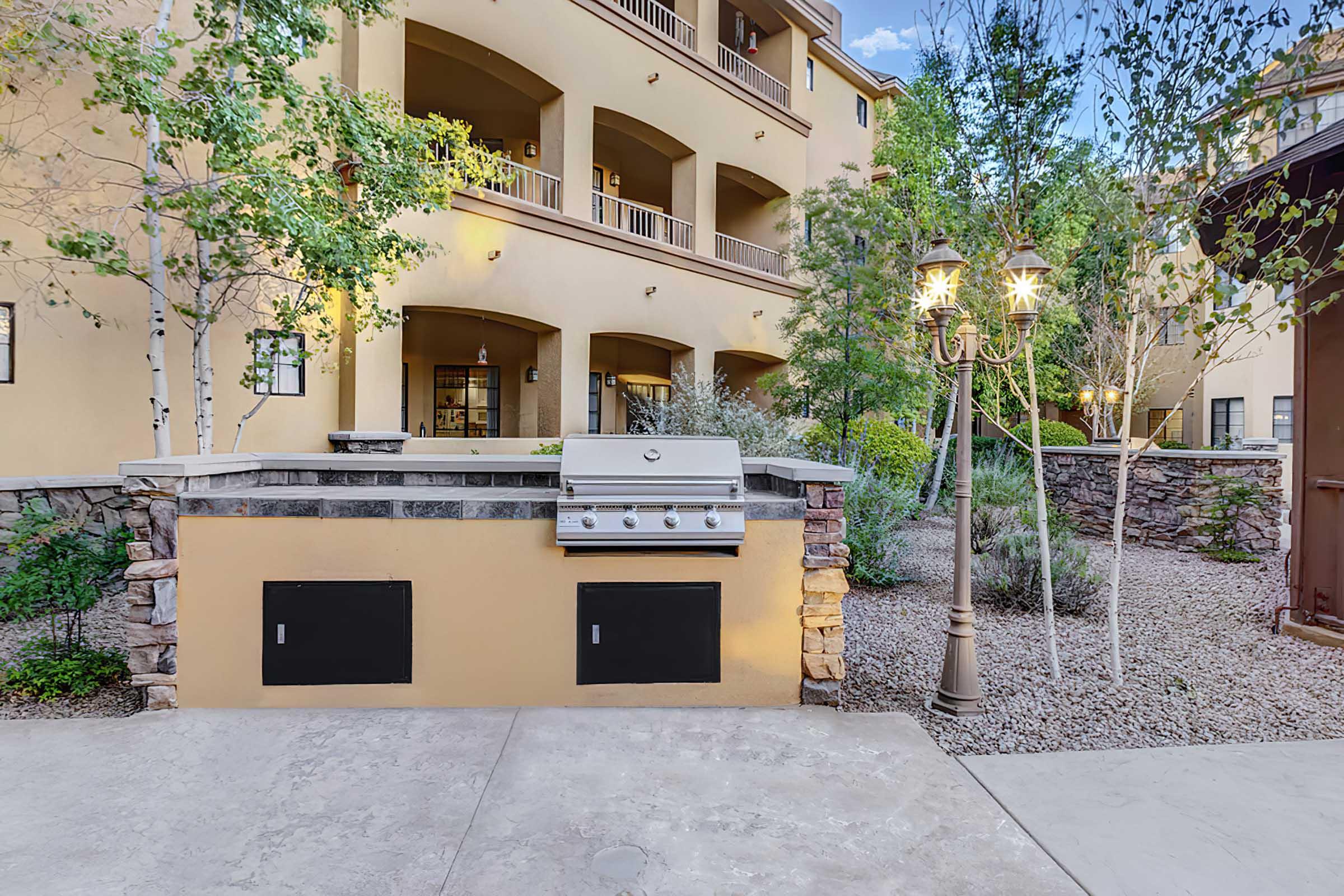 A modern outdoor grill set against a landscaped area with shrubs and trees. The background features a multi-story building with balconies, and a decorative lamp post adds ambiance to the setting. 