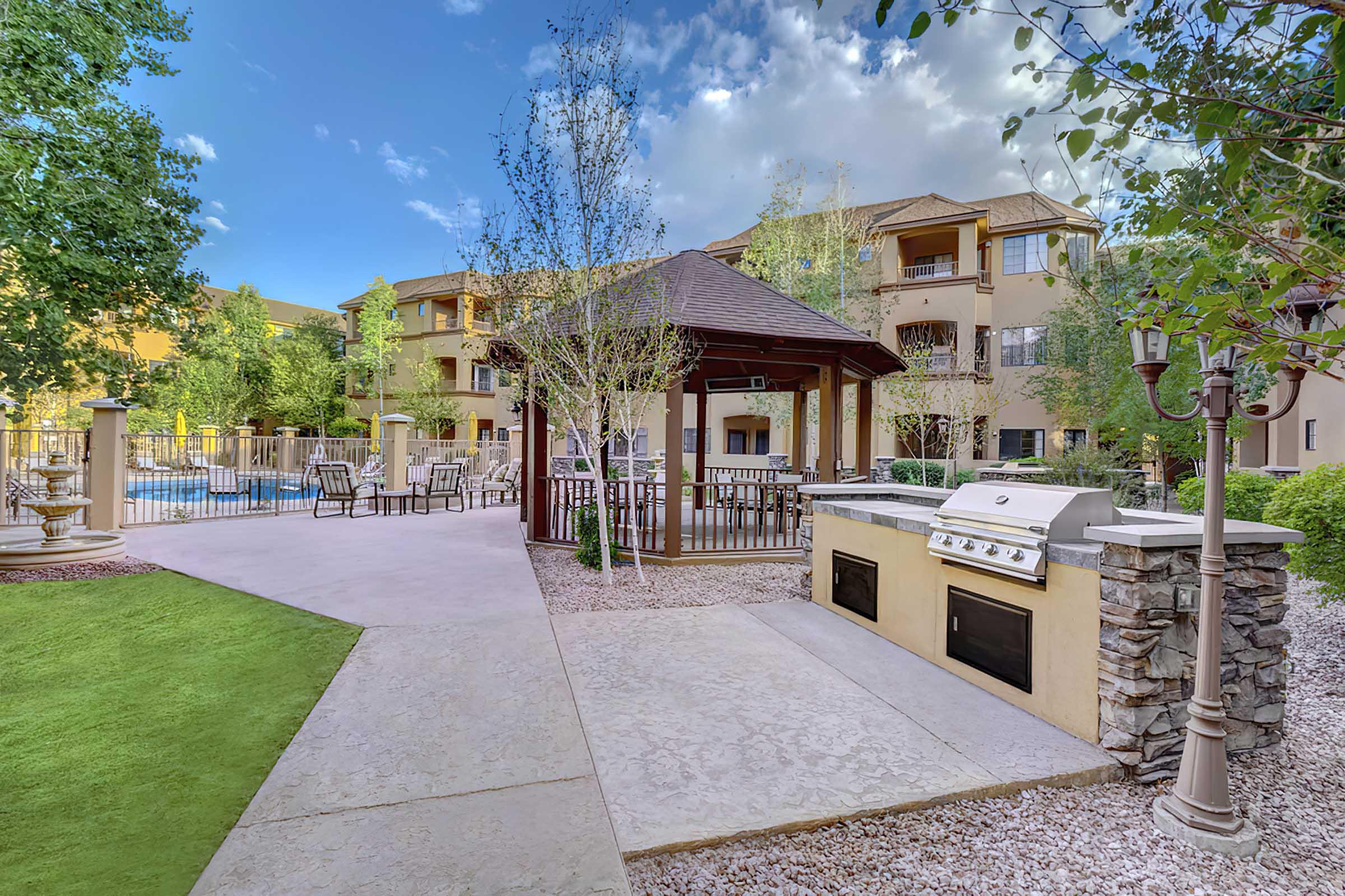 Outdoor area featuring a barbecue grill and seating, surrounded by trees and landscaped grass. In the background, a pool area is visible next to a multi-story building, under a partly cloudy sky.