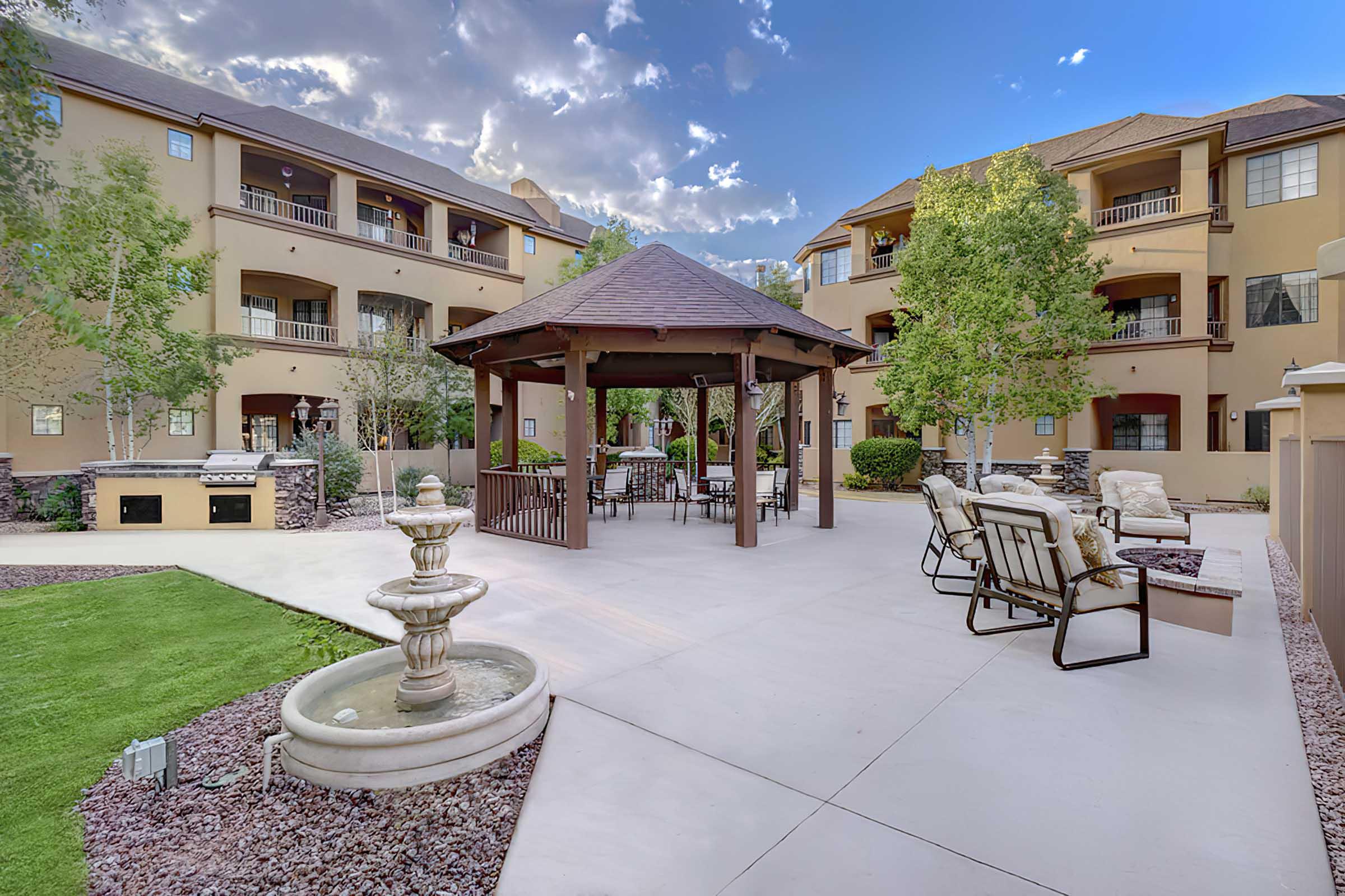 A cozy outdoor courtyard featuring a gazebo with seating, surrounded by landscaped greenery and trees. In the foreground, there's a decorative fountain and comfortable lounge chairs. 