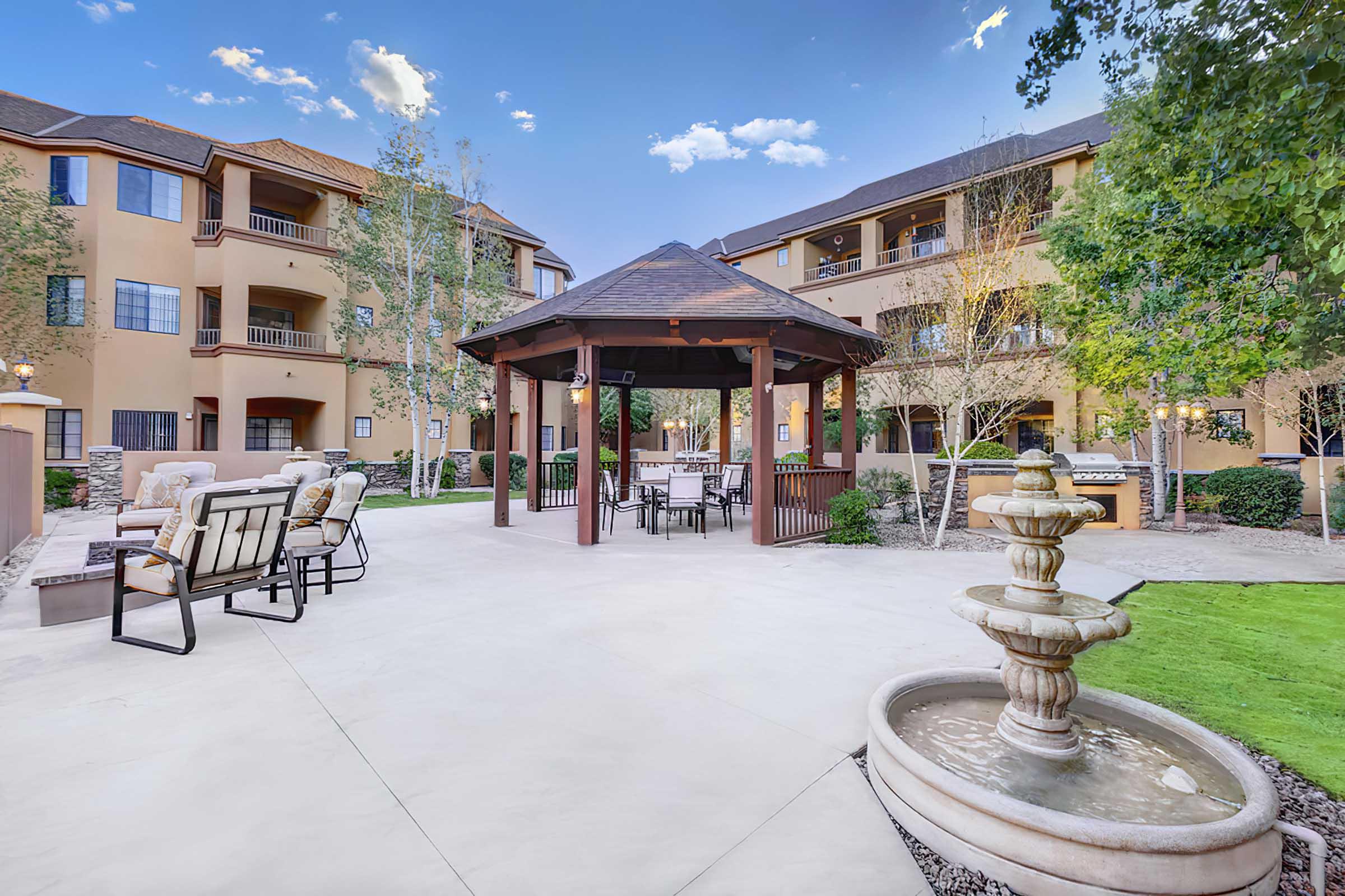 A landscaped courtyard featuring a gazebo with seating, a fountain, and well-maintained greenery, surrounded by residential apartment buildings under a clear blue sky.