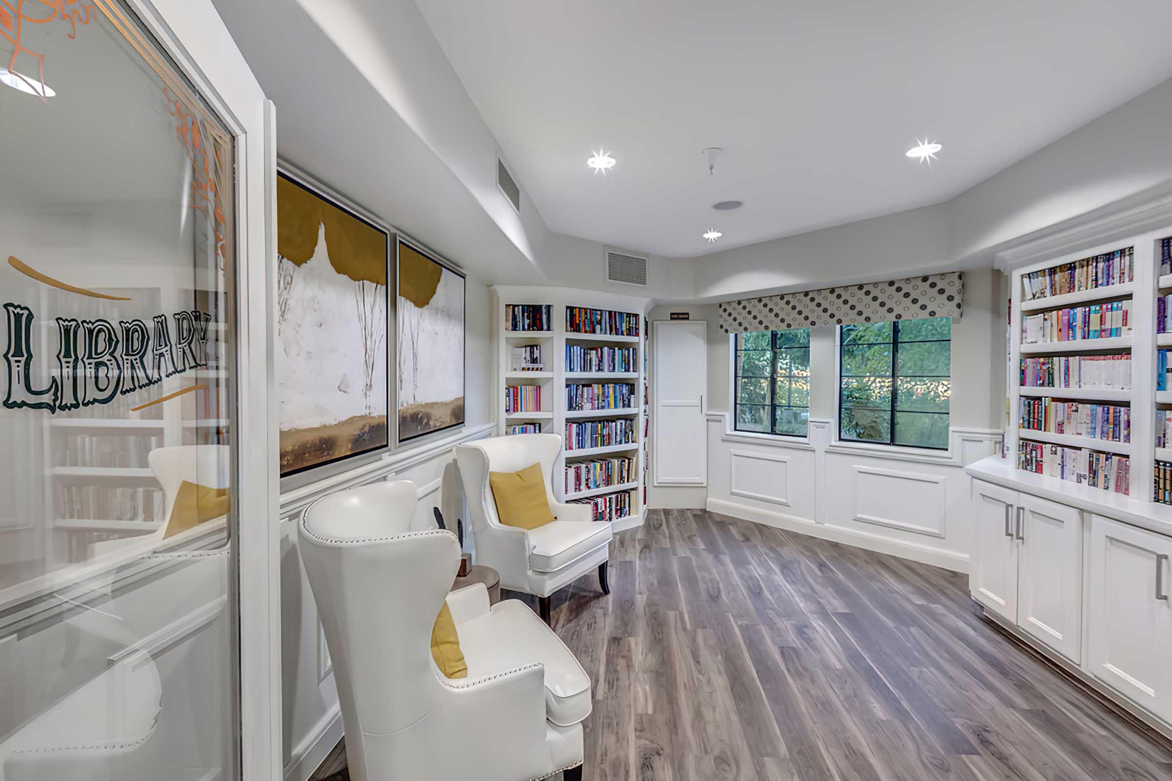 A cozy library room with white walls, large windows letting in natural light, and bookshelves filled with books. 