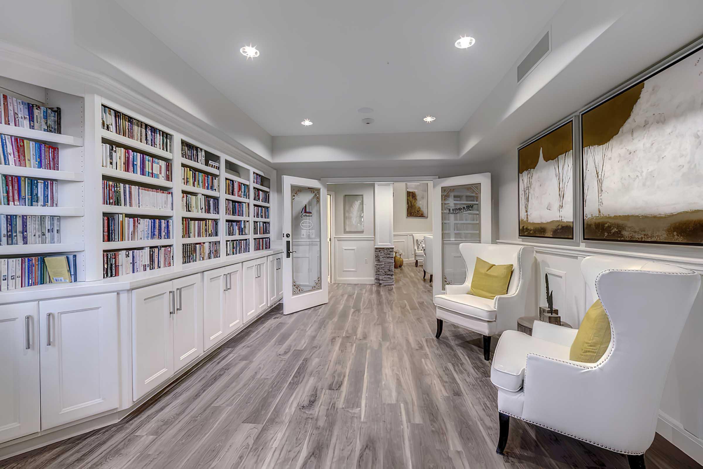A cozy room featuring a white bookshelf filled with books along one wall, two stylish white chairs with yellow cushions, and a decorative art piece. 