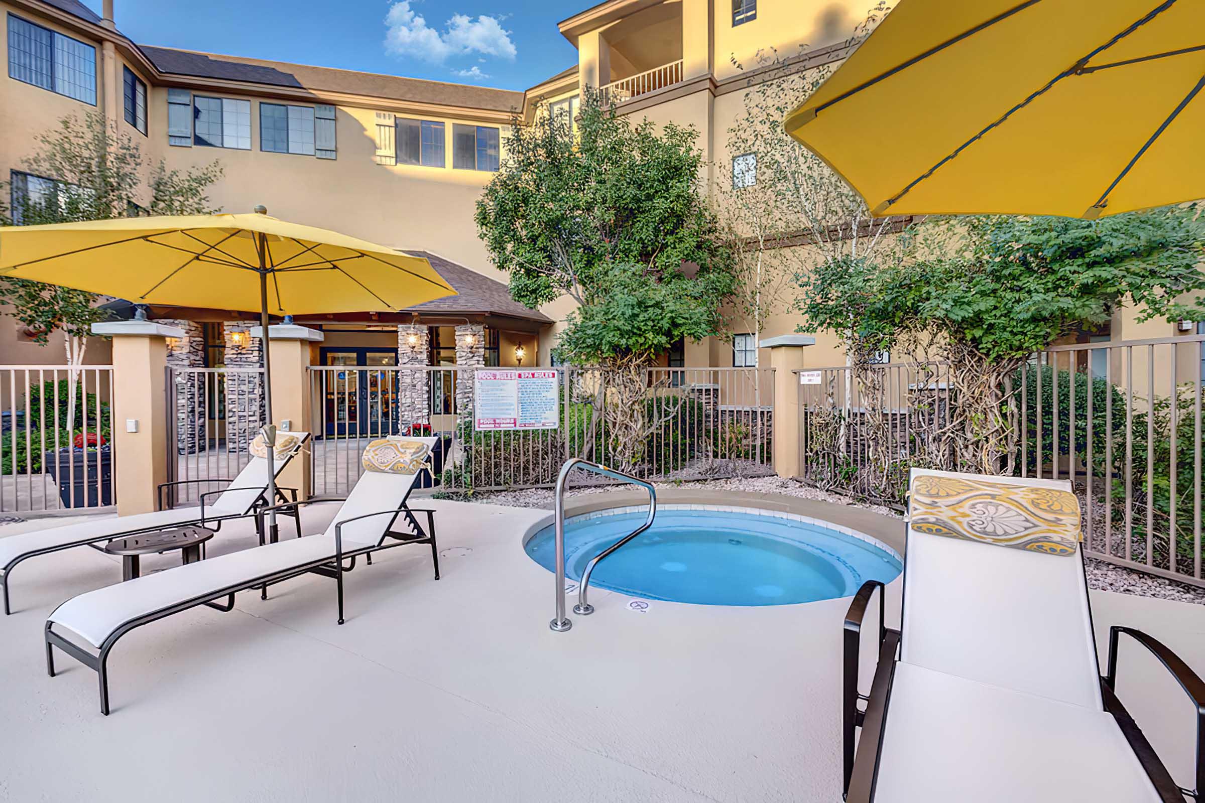 A sunny outdoor pool area features a small hot tub surrounded by lounge chairs and yellow umbrellas. 