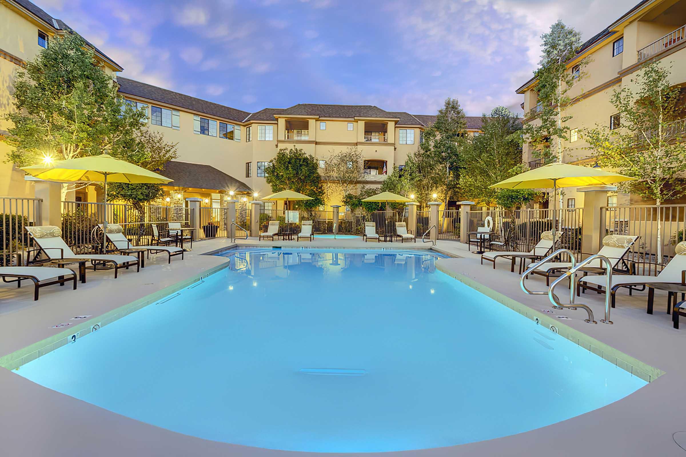 A serene pool area surrounded by lounge chairs and yellow umbrellas, located in a landscaped courtyard of an apartment complex. 