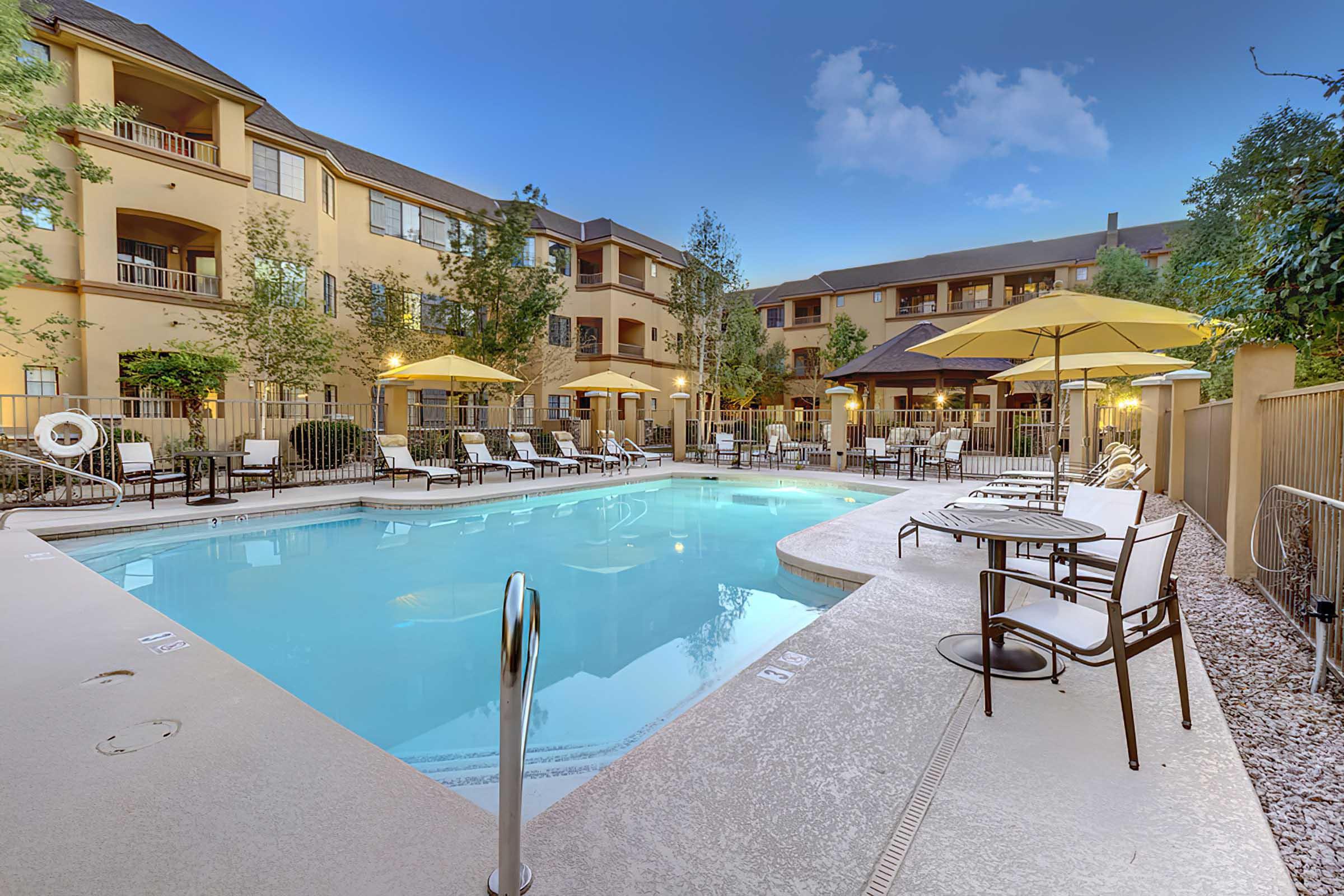 A serene pool area surrounded by apartment buildings. The pool features comfortable lounge chairs, yellow umbrellas, and well-maintained landscaping.