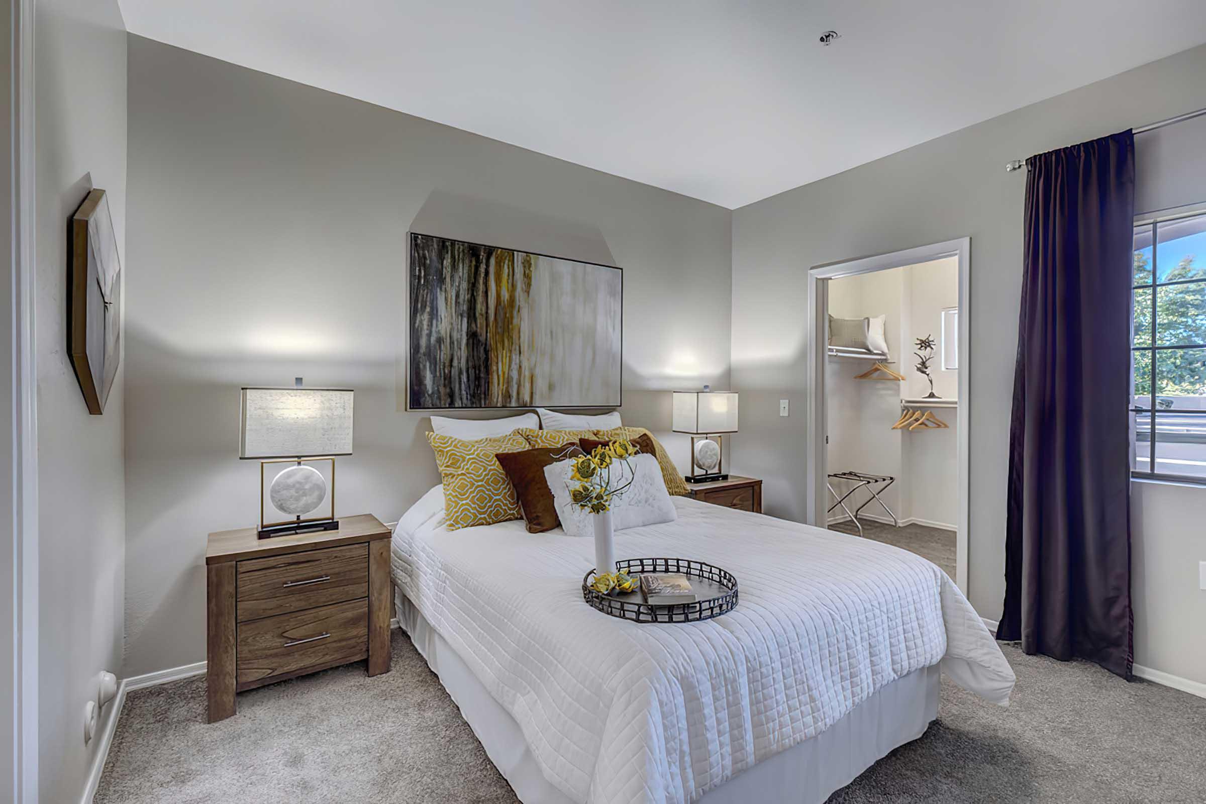 A cozy bedroom featuring a neatly made bed with decorative pillows, two bedside tables with lamps, and a piece of abstract wall art. 