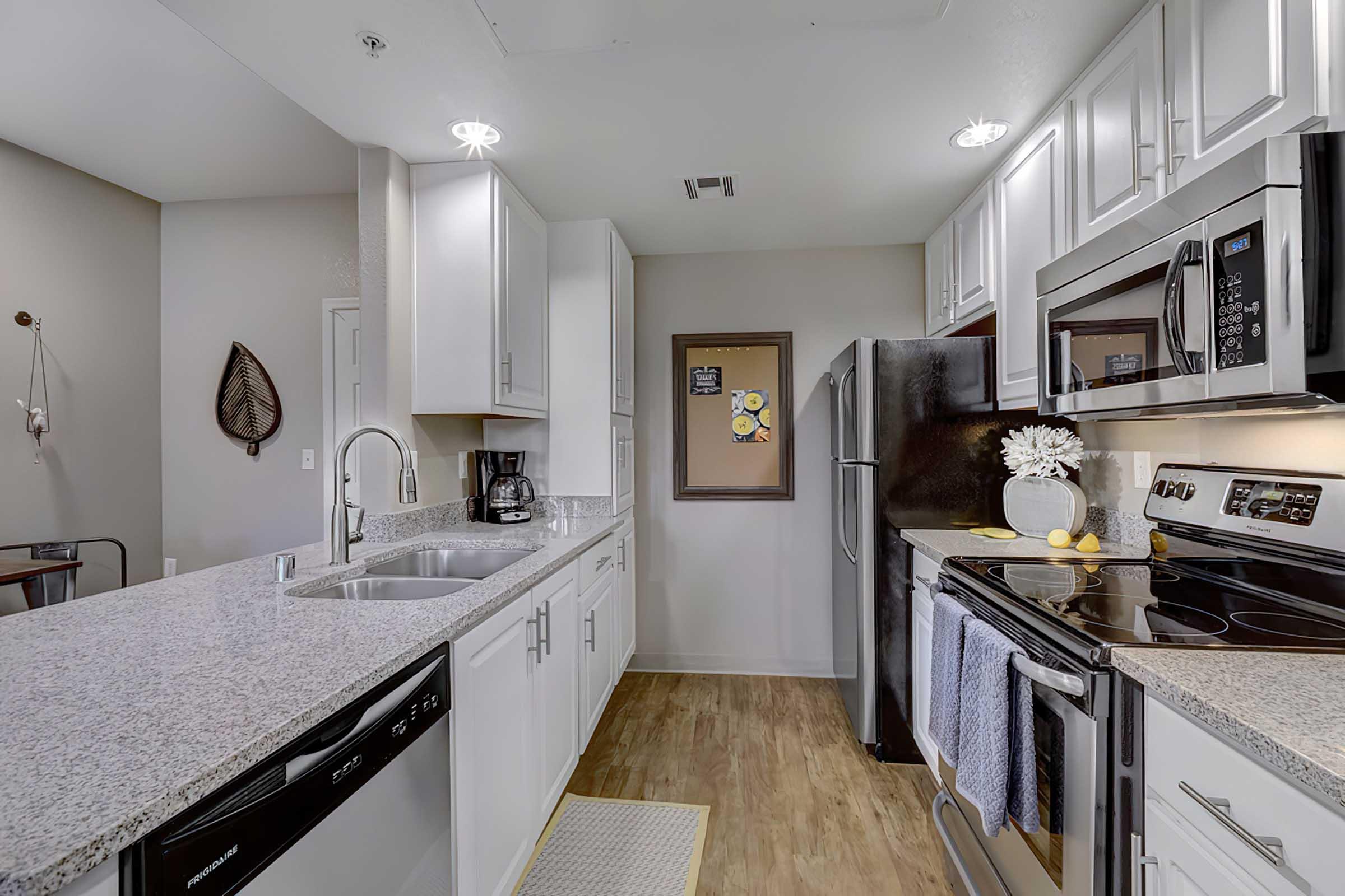 A modern kitchen featuring white cabinetry, stainless steel appliances, and a light granite countertop. It includes a double sink, a microwave, an oven, and a refrigerator