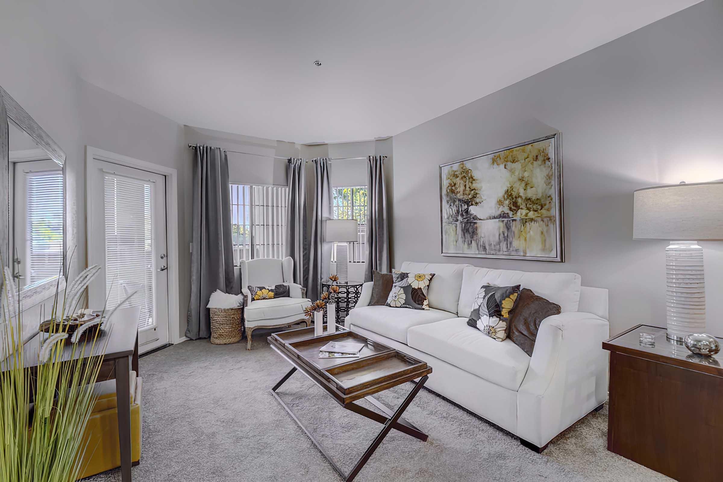 A cozy living room featuring a white sofa with decorative pillows, an accent chair, and large windows with light gray curtains. 