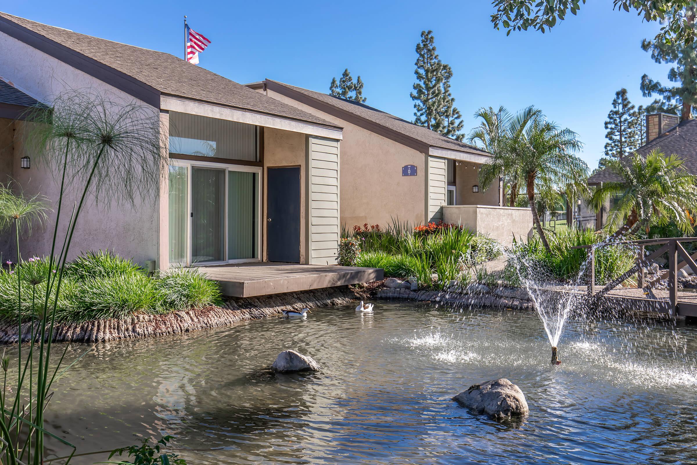A serene landscape featuring a modern building with a porch, surrounded by lush greenery and palm trees. In front, there's a tranquil pond with a fountain and rocks. The scene is brightened by a clear blue sky and an American flag displayed on the building.