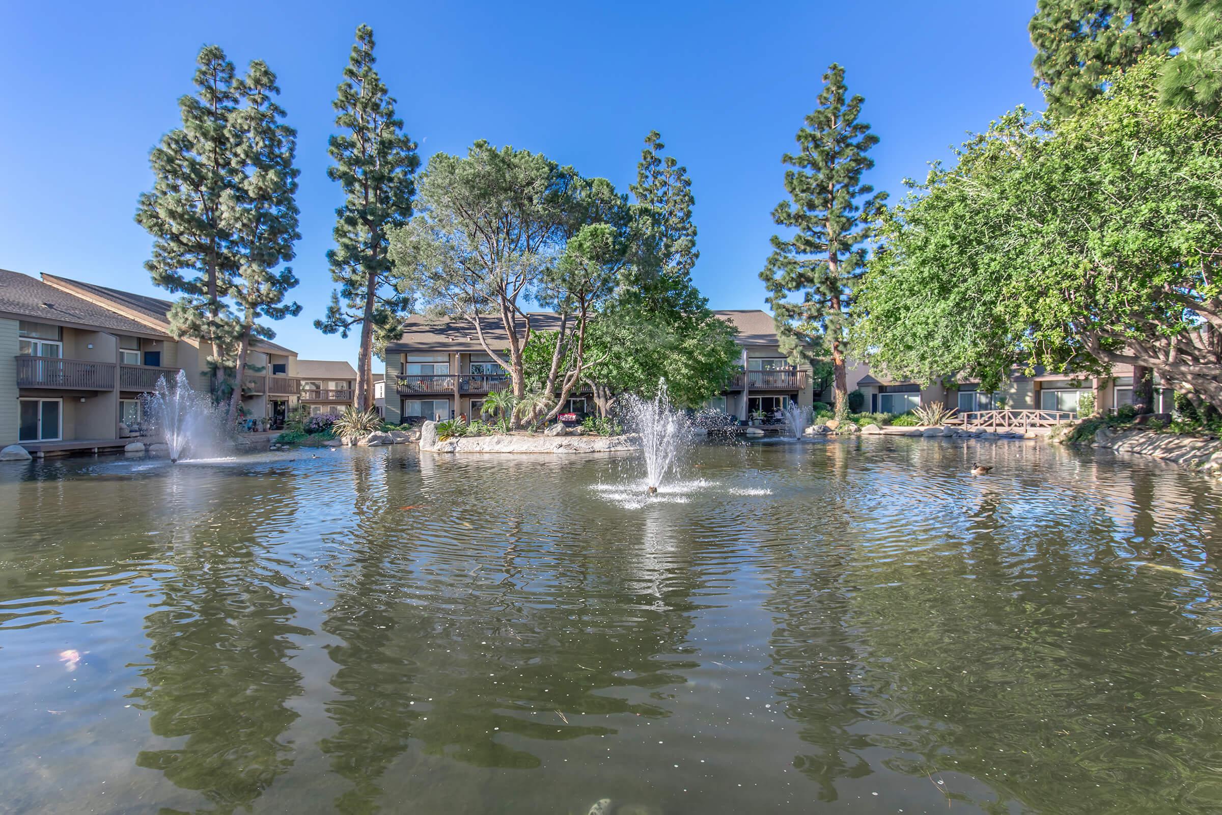 A serene pond surrounded by landscaped trees and residential buildings. Fountains spray water into the air, creating a tranquil atmosphere. The clear blue sky reflects on the water's surface, enhancing the peaceful setting.