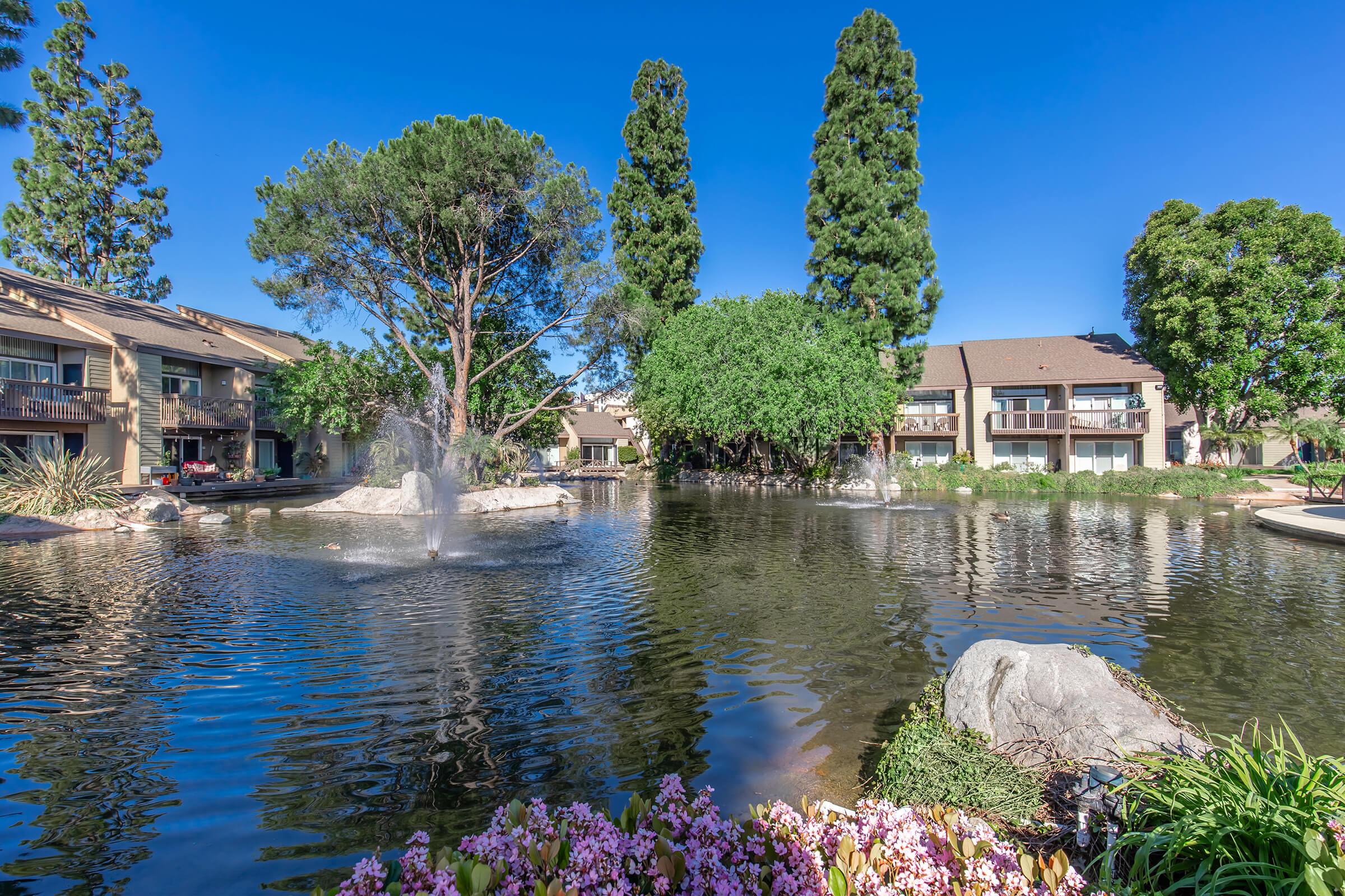 A serene pond surrounded by lush greenery and blooming flowers. Water fountains create gentle ripples in the water, reflecting the clear blue sky. In the background, there are residential buildings nestled among tall trees, creating a peaceful, nature-inspired setting.