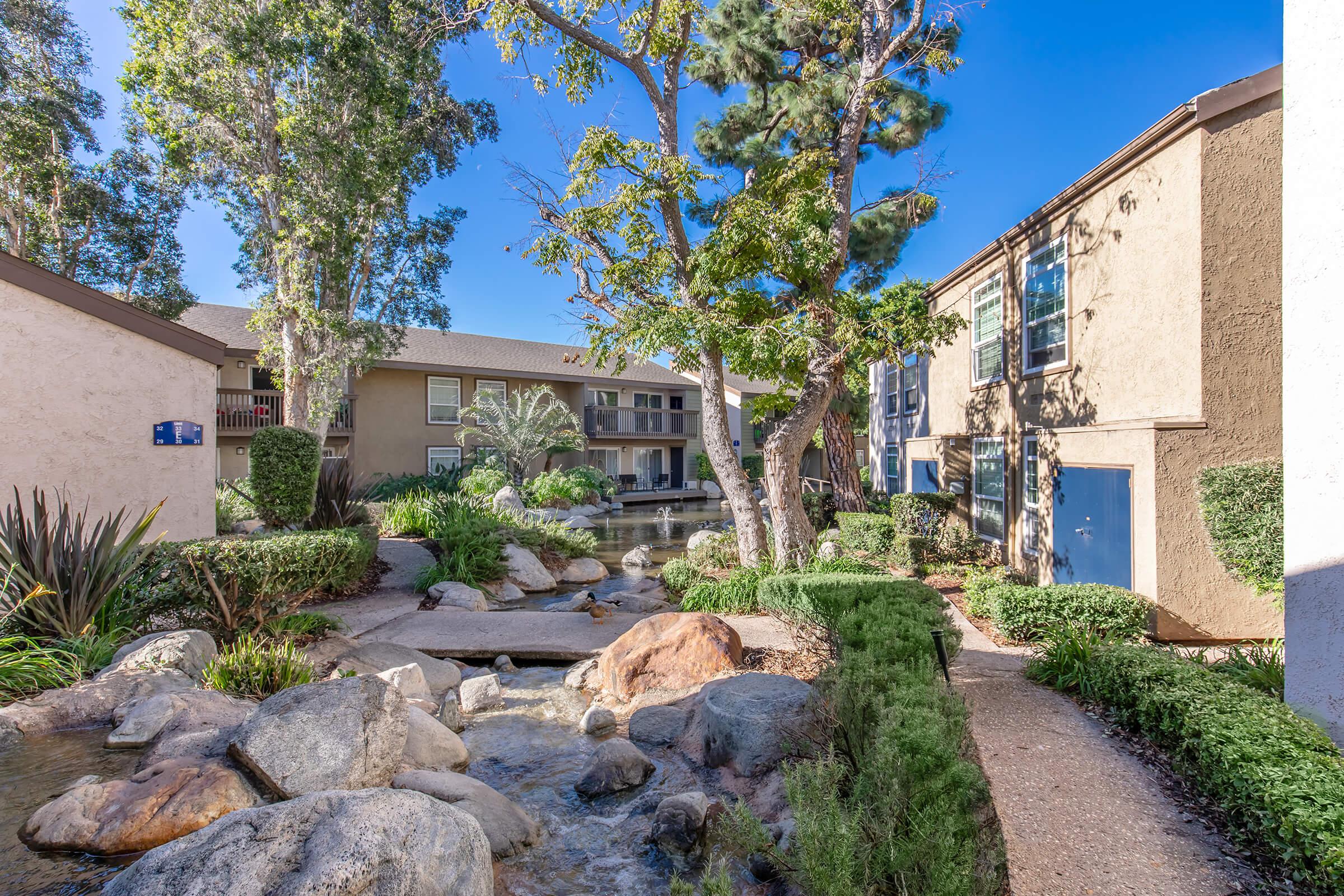 A tranquil scene of an apartment complex featuring a landscaped pathway with rocks and shrubs, a pond surrounded by trees, and two buildings in the background under a clear blue sky. The area exudes a peaceful and inviting atmosphere.