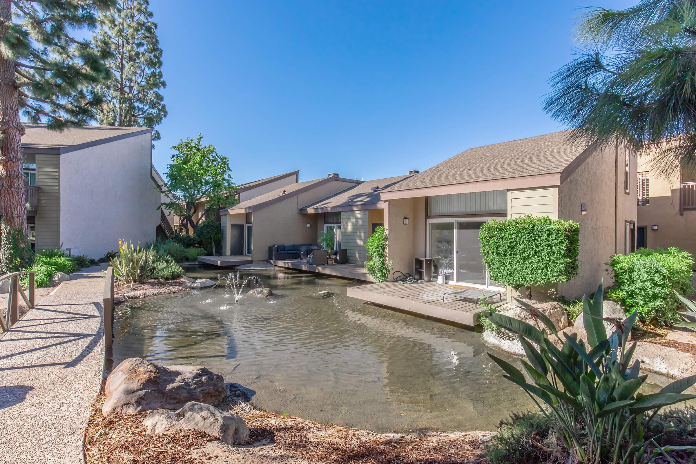 Tranquil outdoor scene featuring modern residential units with neutral-colored exteriors. A small pond with fountains and surrounding greenery enhances the peaceful ambiance. The pathway leads to a cozy seating area, inviting relaxation in a serene environment.