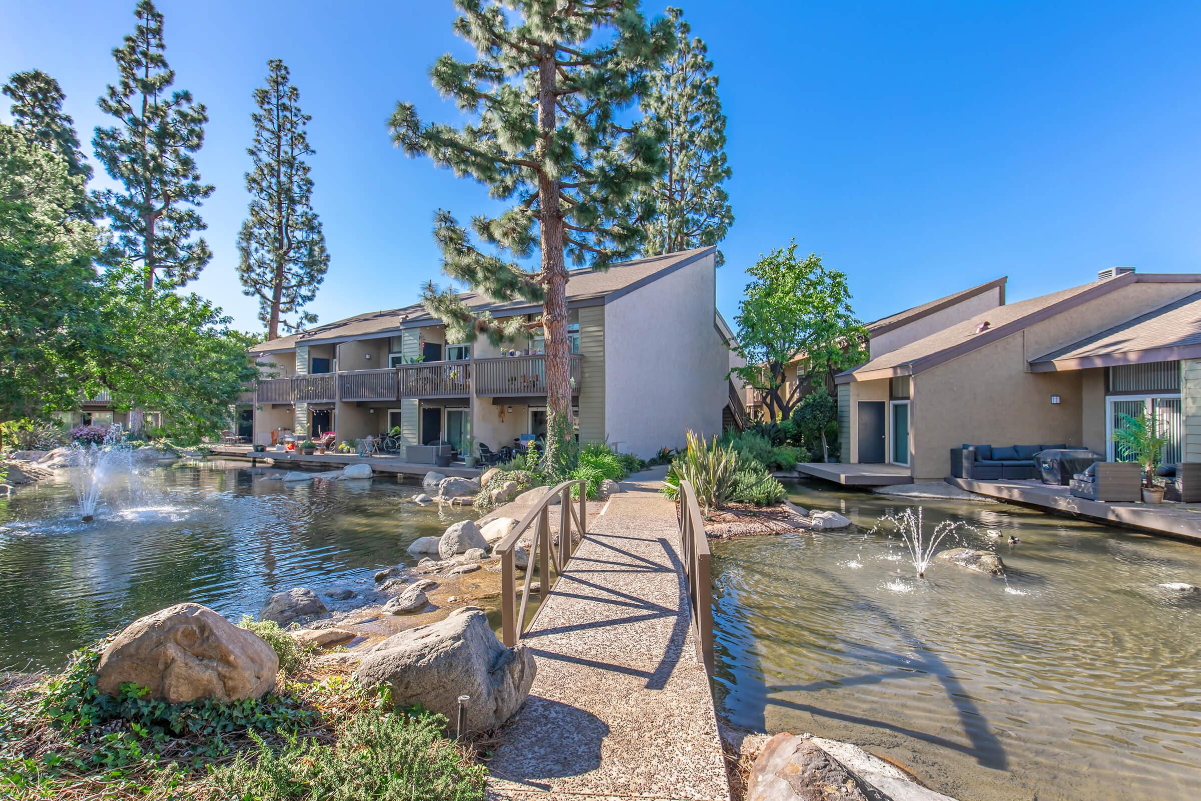 A scenic view of a residential complex surrounded by greenery, featuring a pond with fountains and a small bridge leading over the water. The buildings have modern architecture and outdoor seating areas, with tall trees in the background and a clear blue sky.