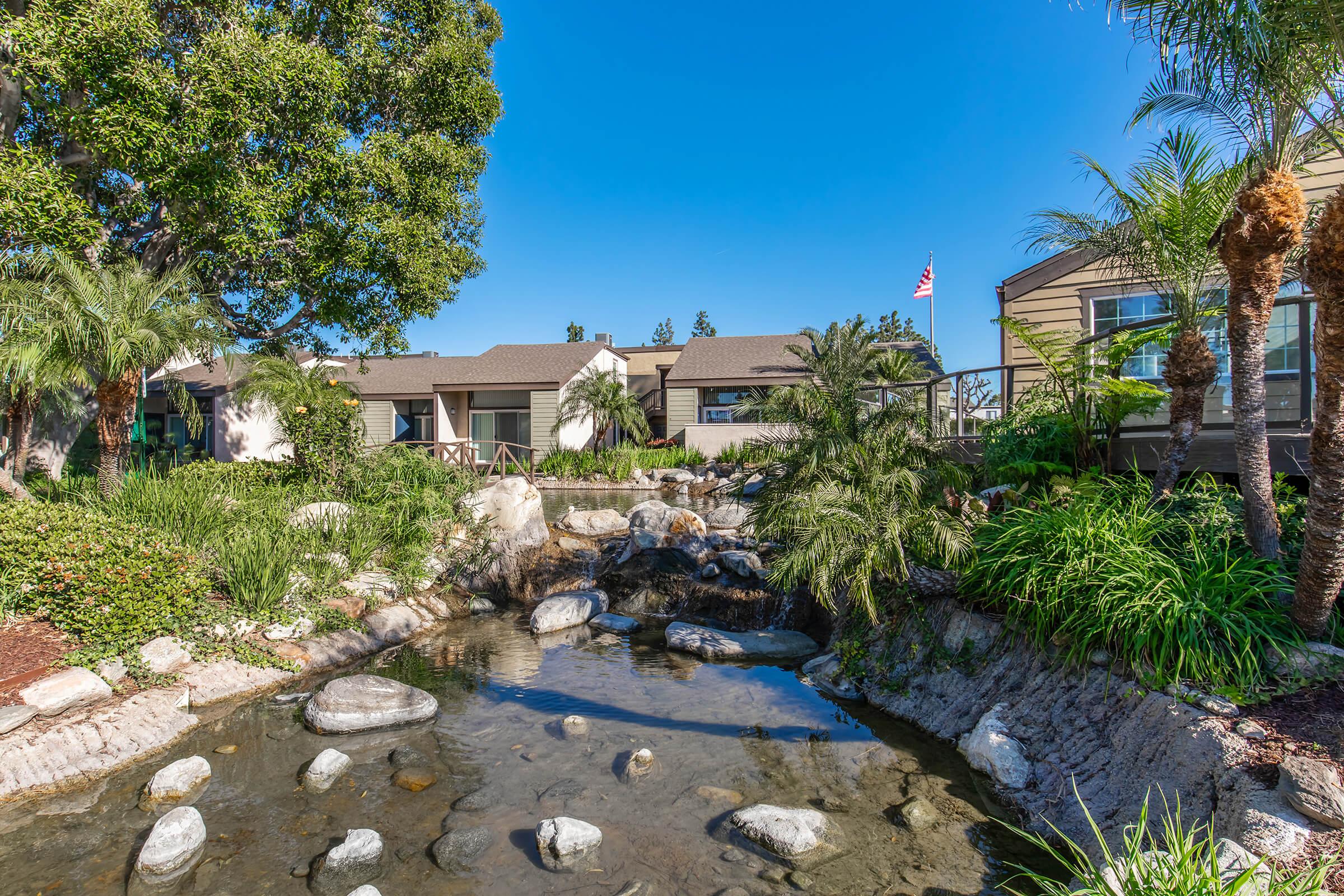 A serene landscape featuring a small pond with rocks and lush greenery, surrounded by palm trees. In the background, there are buildings with brown roofs and a clear blue sky. A flag is visible, adding a touch of color to the tranquil scene.
