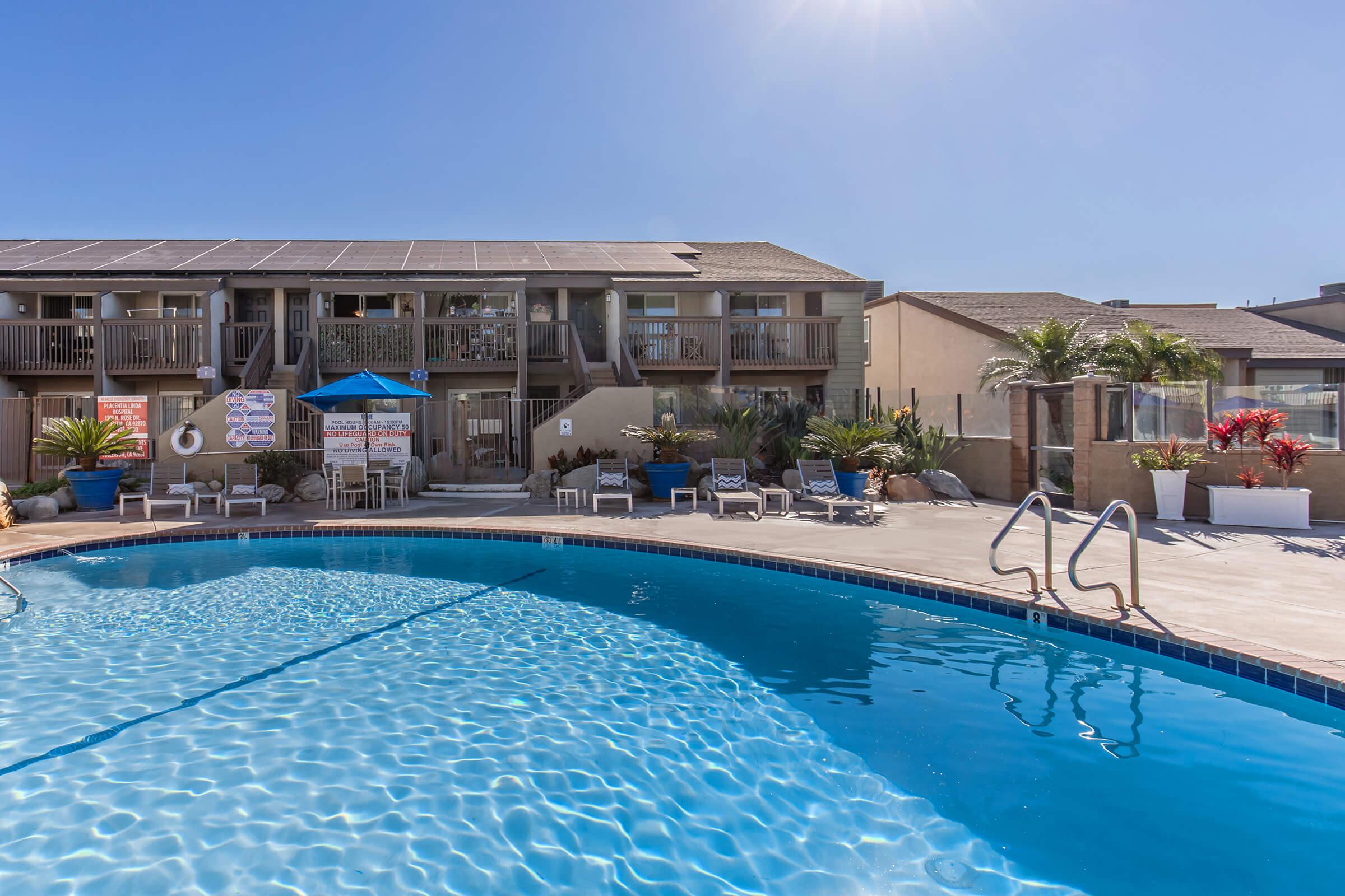 Swimming pool with clear blue water, surrounded by lounge chairs and potted plants. In the background, a two-story building features balconies, tables with umbrellas, and signs. Bright sunlight illuminates the scene, creating a relaxing atmosphere.