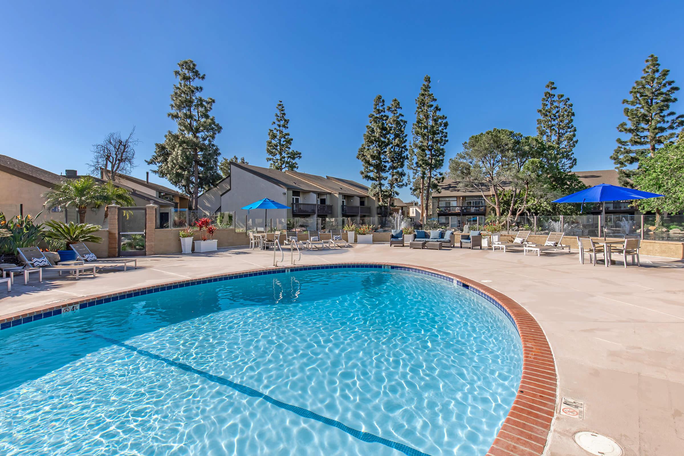 A serene outdoor swimming pool surrounded by sun loungers and umbrellas, with tall trees in the background and residential buildings nearby. The clear blue water reflects the bright blue sky, creating a relaxing atmosphere perfect for leisure and enjoyment.