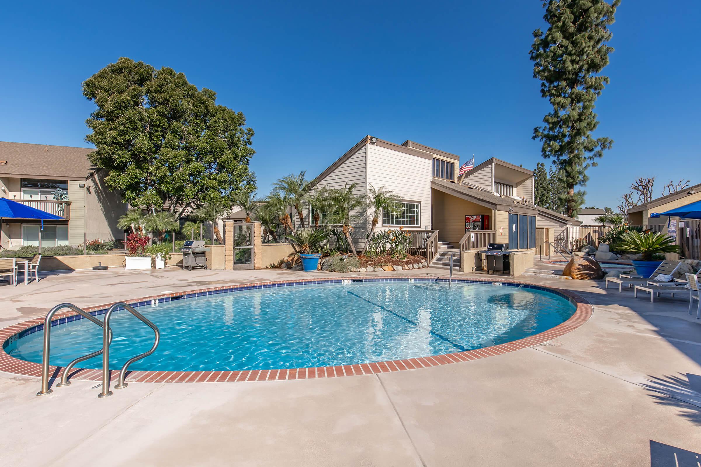 Swimming pool surrounded by lounge chairs and palm trees, with a residential building in the background. Clear blue sky overhead, creating a bright and inviting atmosphere. The area features landscaped greenery and outdoor amenities, perfect for relaxation and leisure.