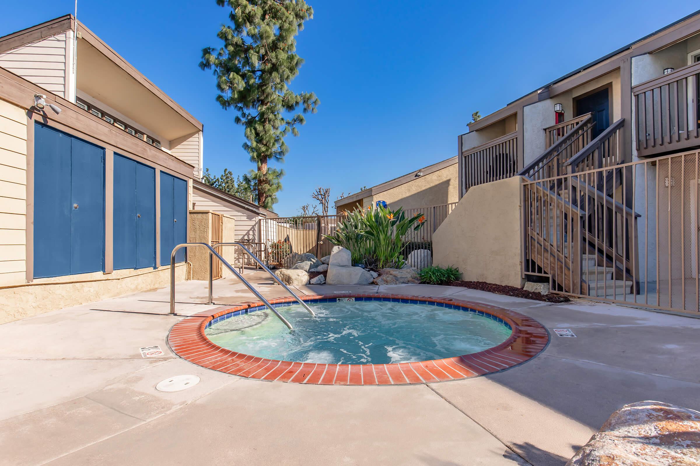 Hot tub surrounded by a paved patio in a residential area. There are stairs leading to upper-level units and a green plant arrangement nearby. Blue storage doors are visible on the left side, under clear blue skies. The setting is well-lit and inviting, suggesting a relaxing outdoor space.