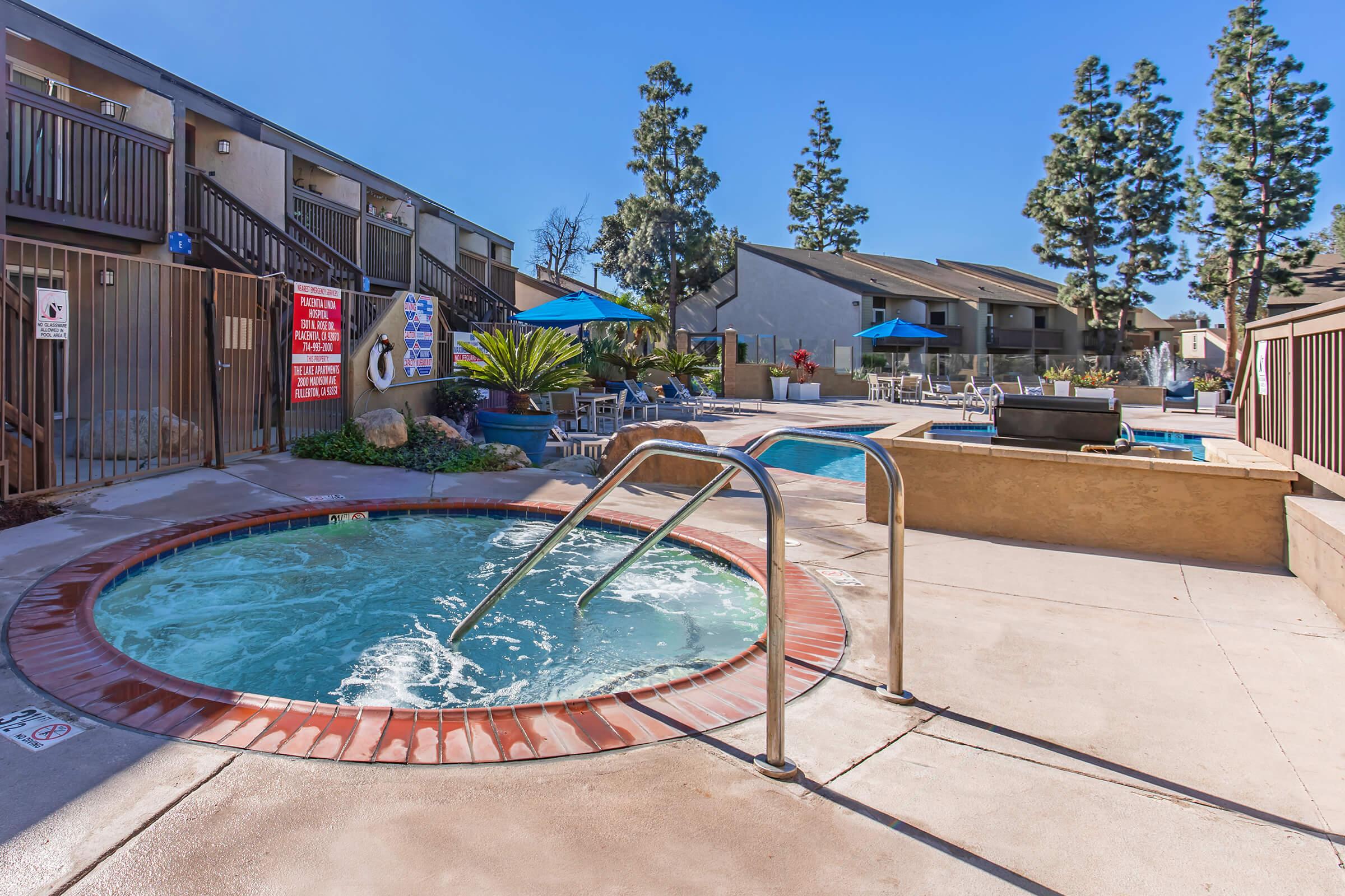 A spacious outdoor area featuring a hot tub with steps, surrounded by palm trees and lounge chairs. In the background, there are swimming pools and several buildings with balconies. Bright blue skies complete the inviting atmosphere.