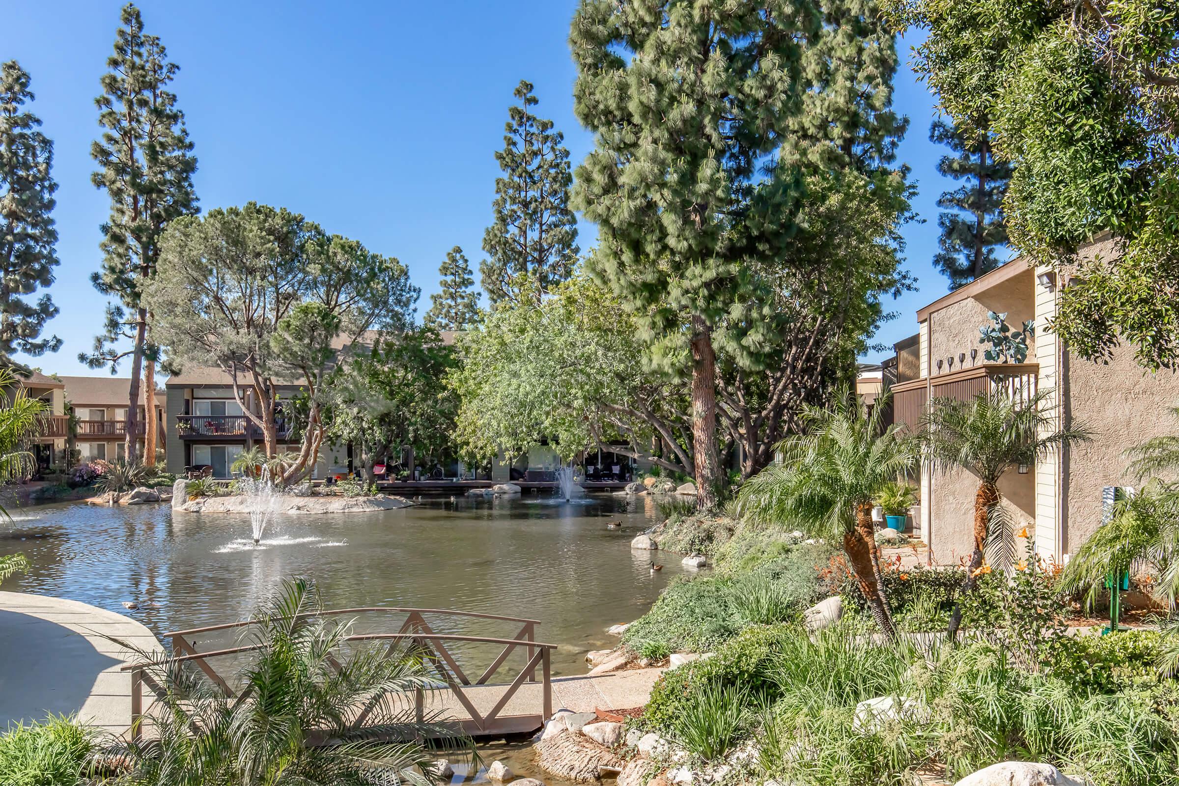 A serene landscape featuring a calm pond surrounded by lush greenery and tall trees. A small wooden bridge crosses the pond, with fountains creating gentle water sprays. Buildings are visible in the background, hinting at a residential setting. The scene is bright and inviting under a clear blue sky.